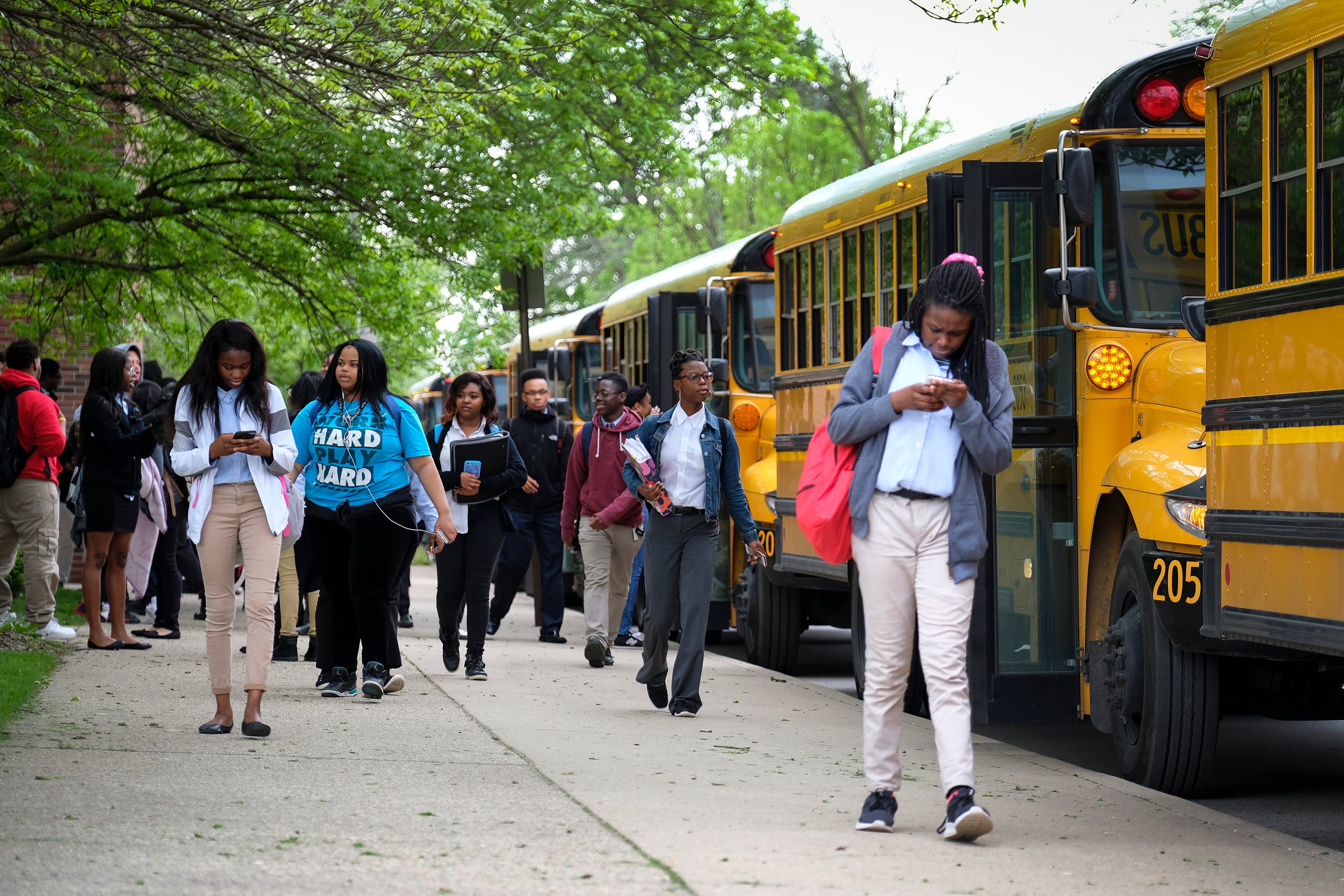 Students walk by a couple of yellow and black school busses with green trees in the background.