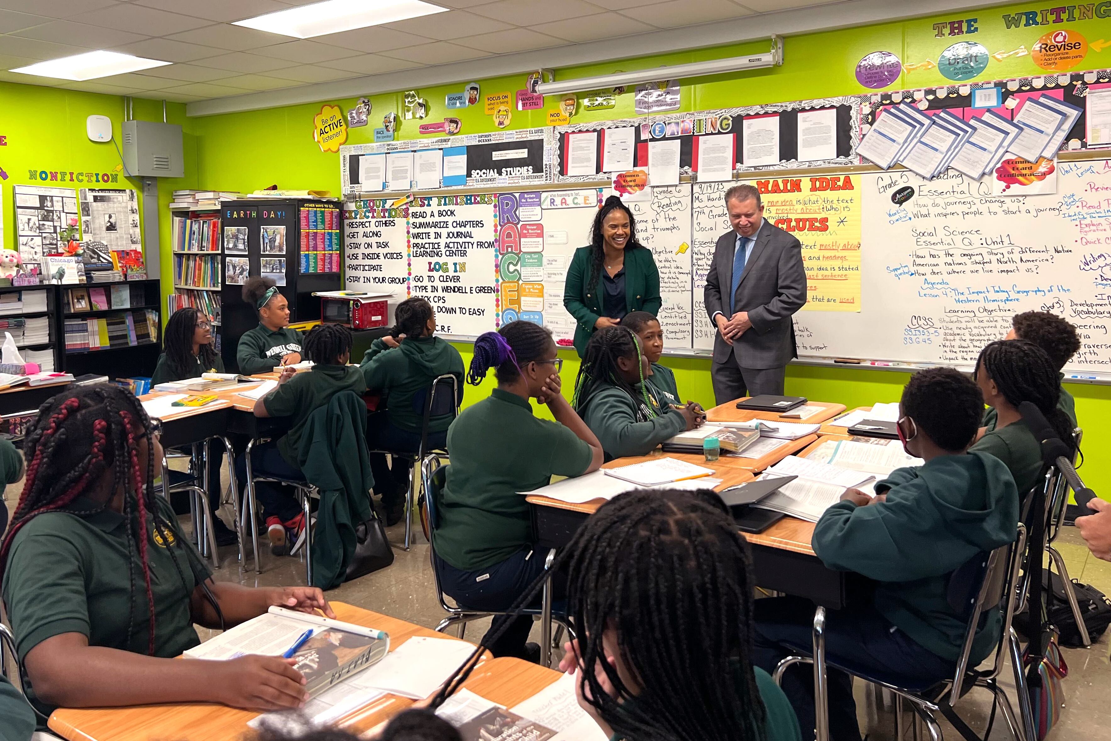 Two adults stand at the front of a classroom full of students sitting at desks.