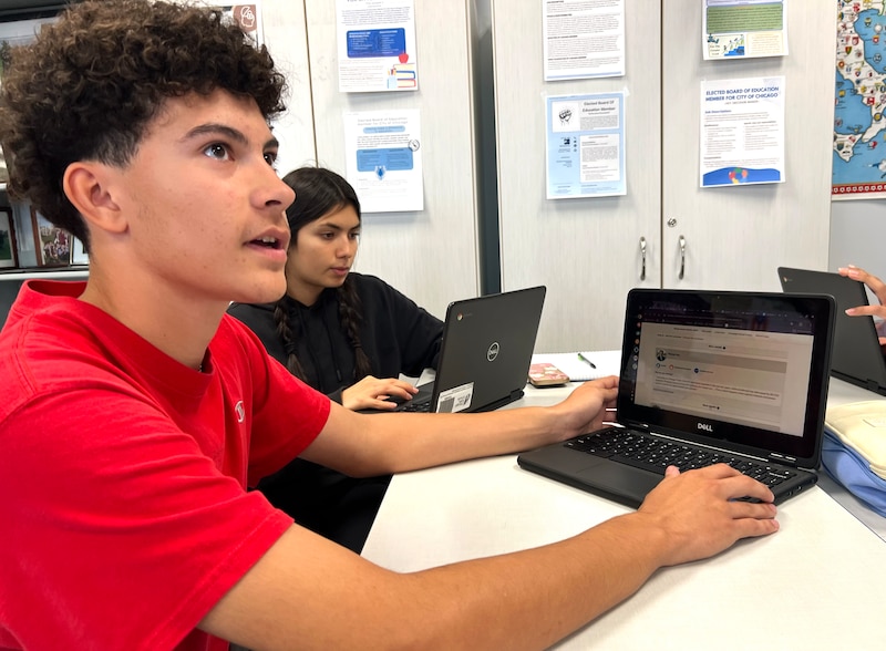 A male student in a red shirt looks to the right in front of a laptop.