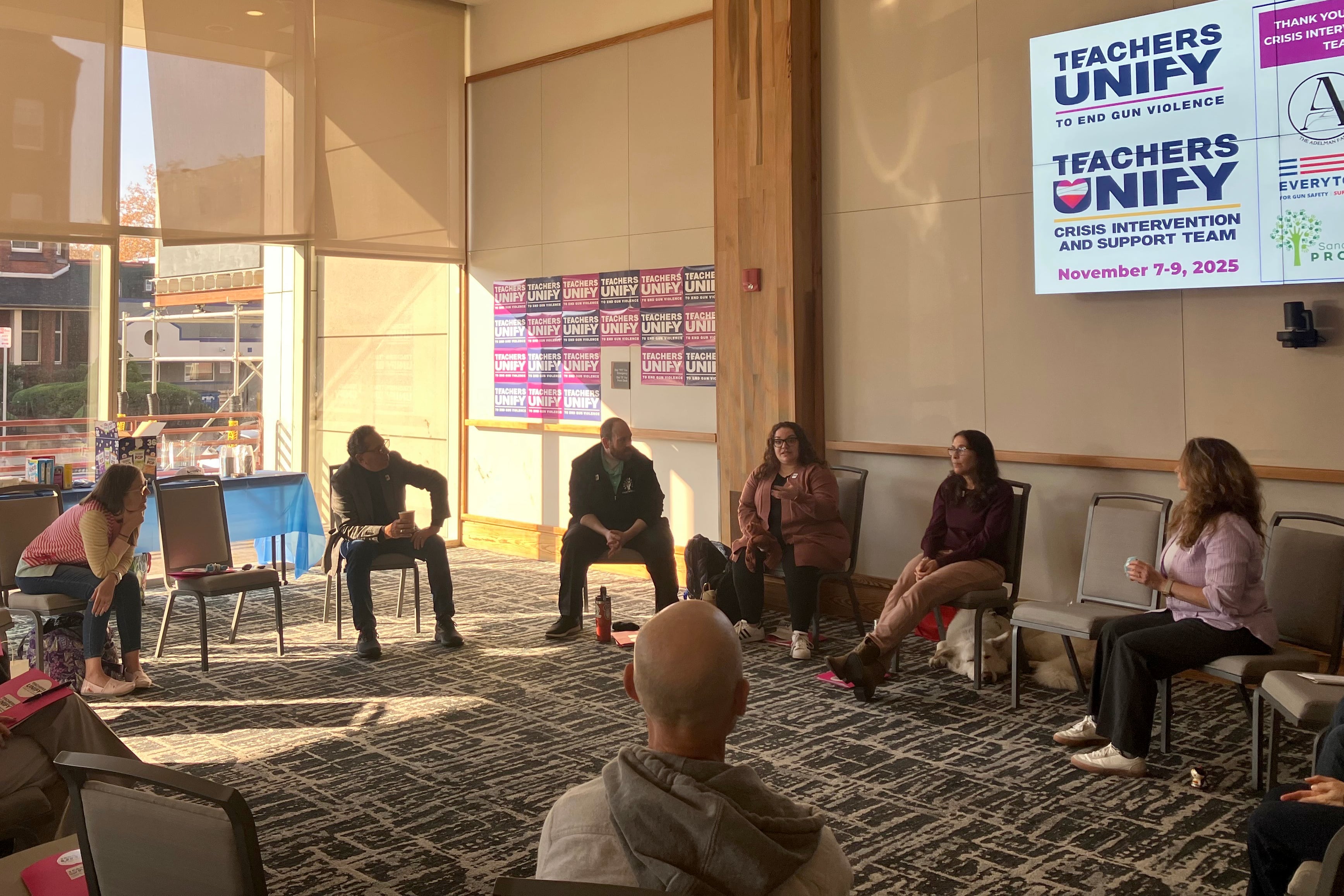 A photograph of a group of people sitting in chairs in a circle in a hotel conference room.