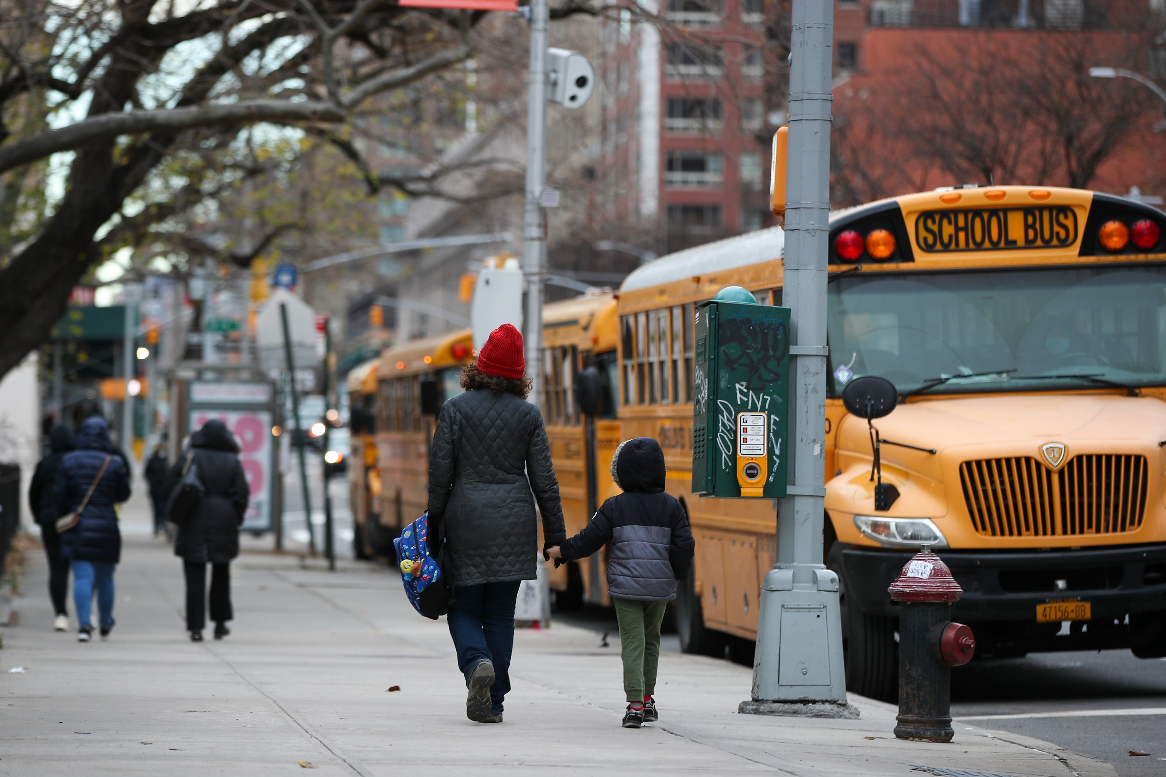 An adult holds the hand of a young child next to a row of yellow school buses. They are all wearing cold clothing.