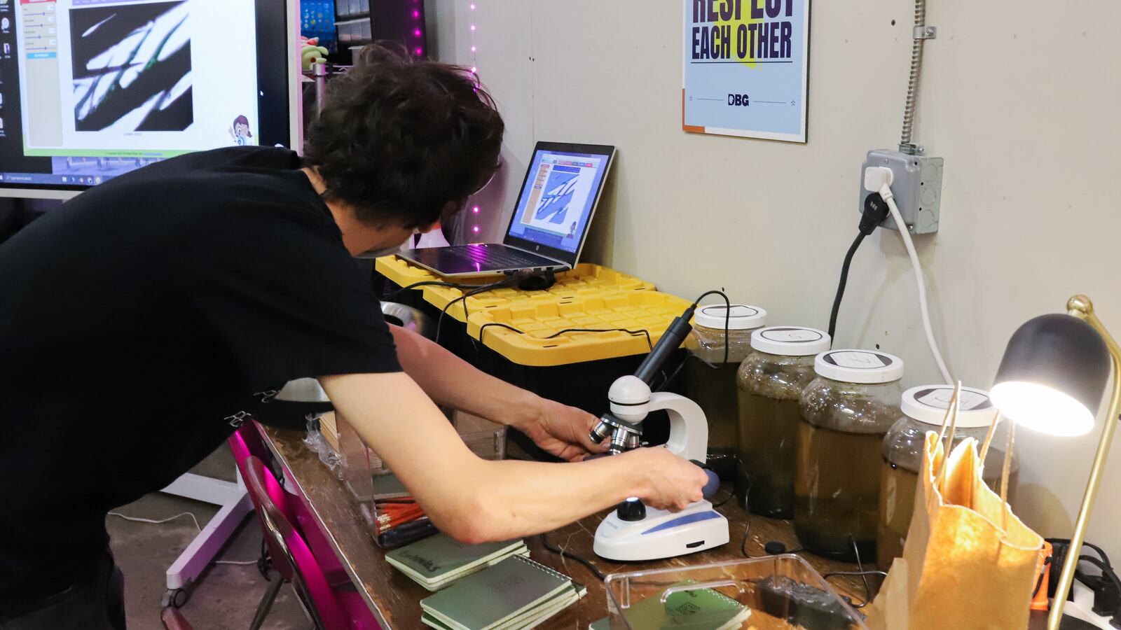 A teacher examines seeds underneath a microscope in a classroom, projecting them onto a computer.