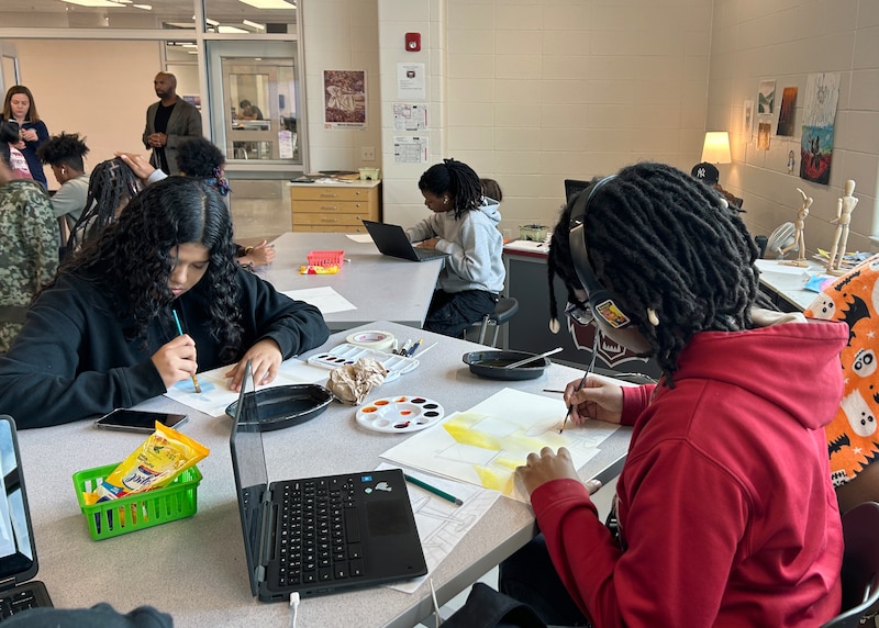 Two high school students sit at a table working on an art project while other students sit at tables in the background and two adults stand near the doorway in the background.
