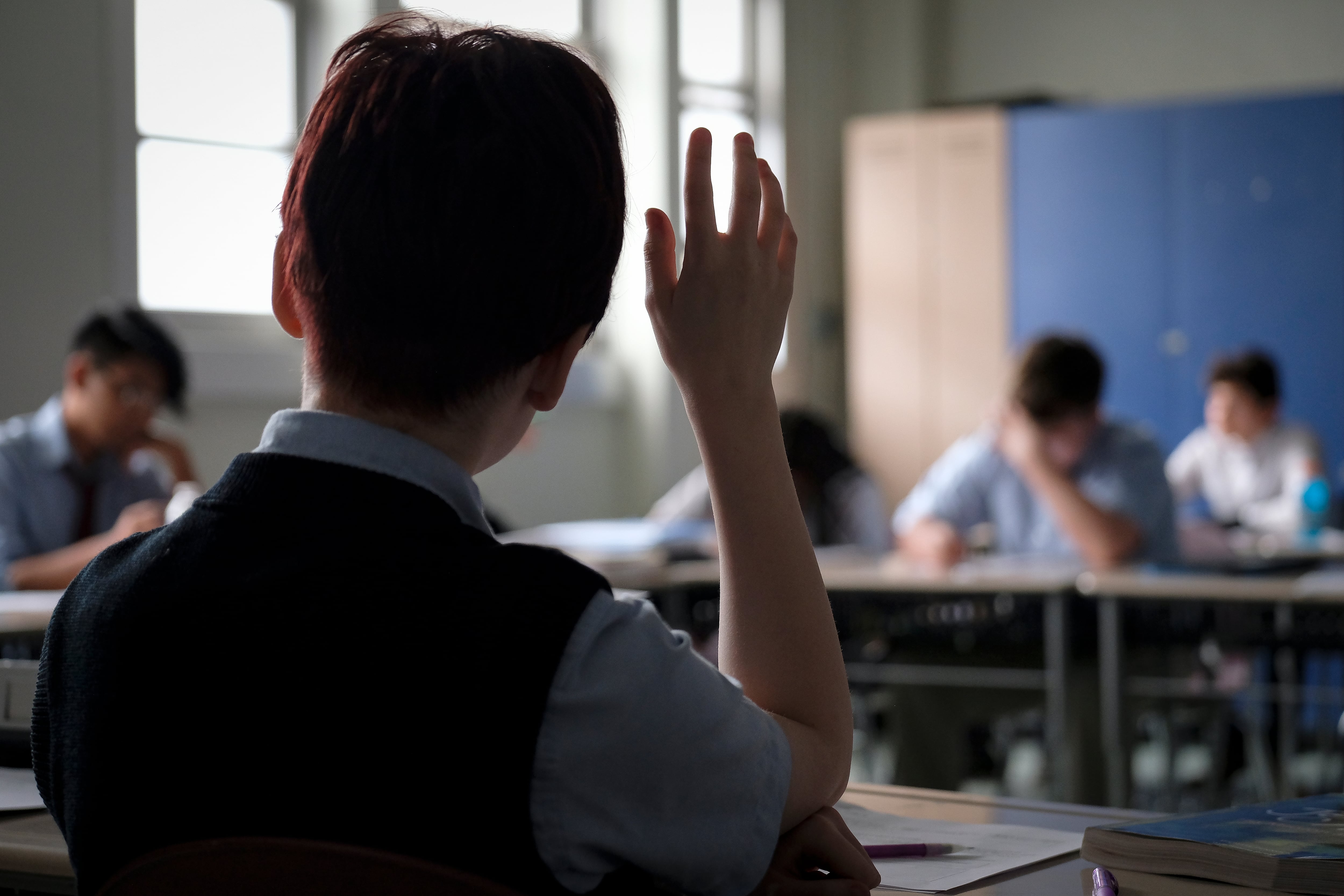 A view of a classroom from behind a student who is raising their hand.