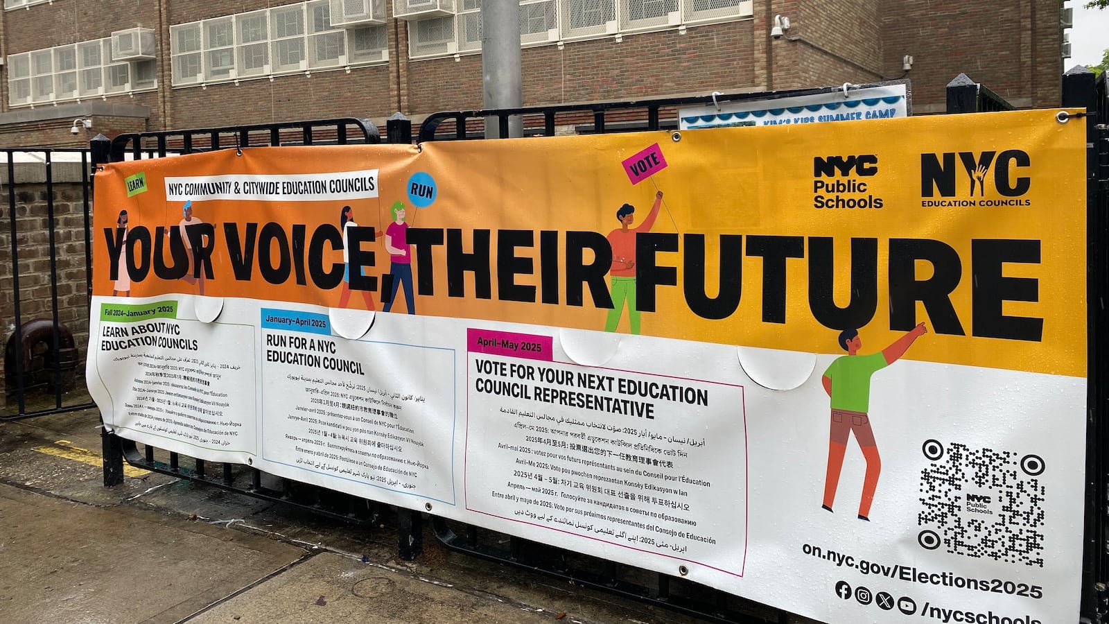 A banner saying "Your voice their future" hangs on a fence in front of a beige brick school building.