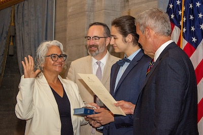 A woman, surrounded by three men, raises one hand, and places her other hand on a Bible.