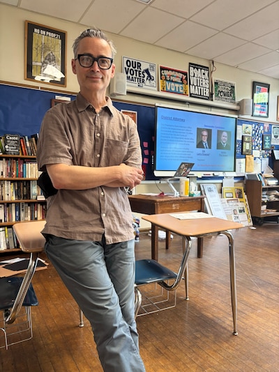 A photograph of a male teacher posing for a photograph while sitting at a desk in a classroom.