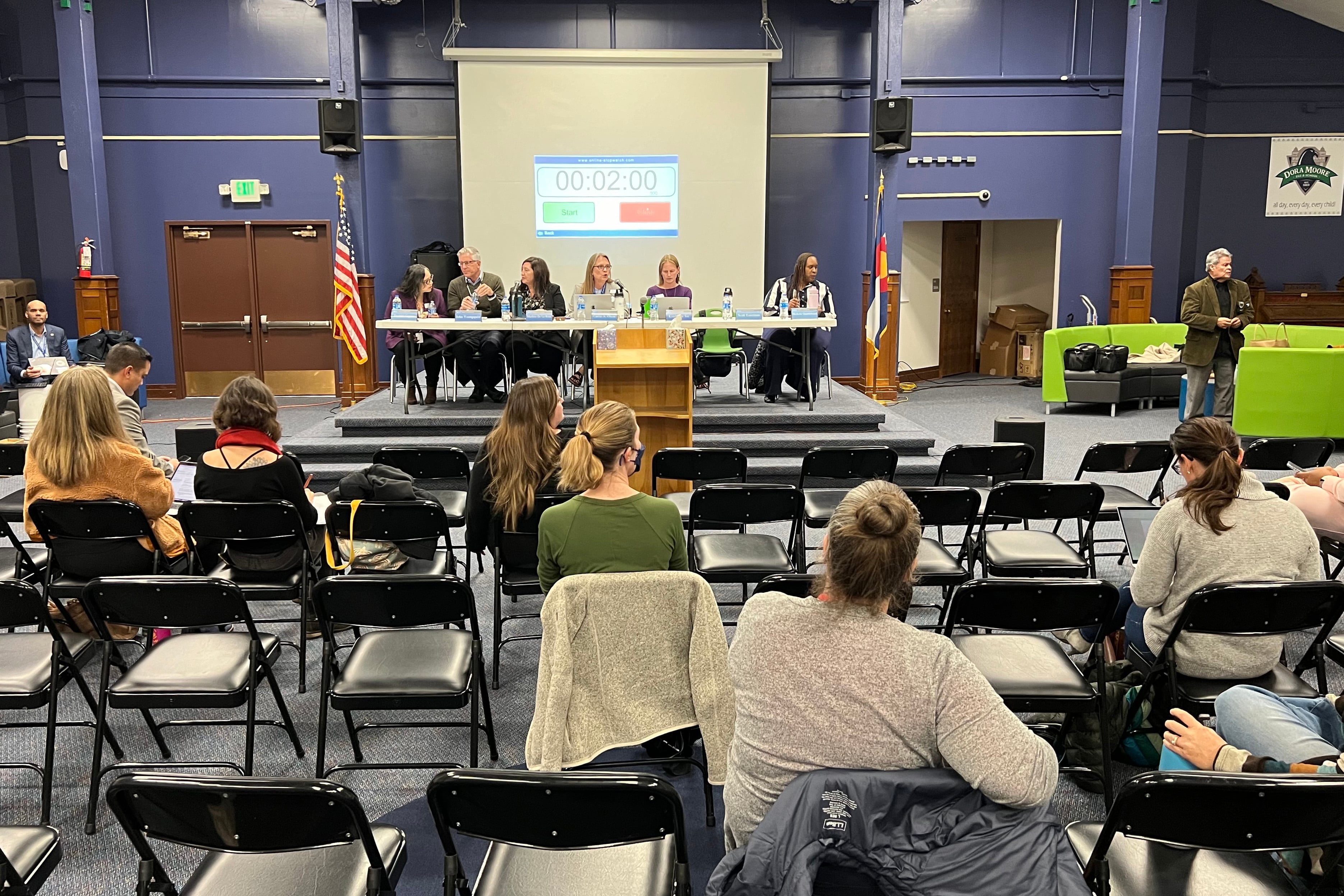 A group of people sit in a row of chairs and tables in the front of a large room with a group of people sitting in chairs facing the people in the front.
