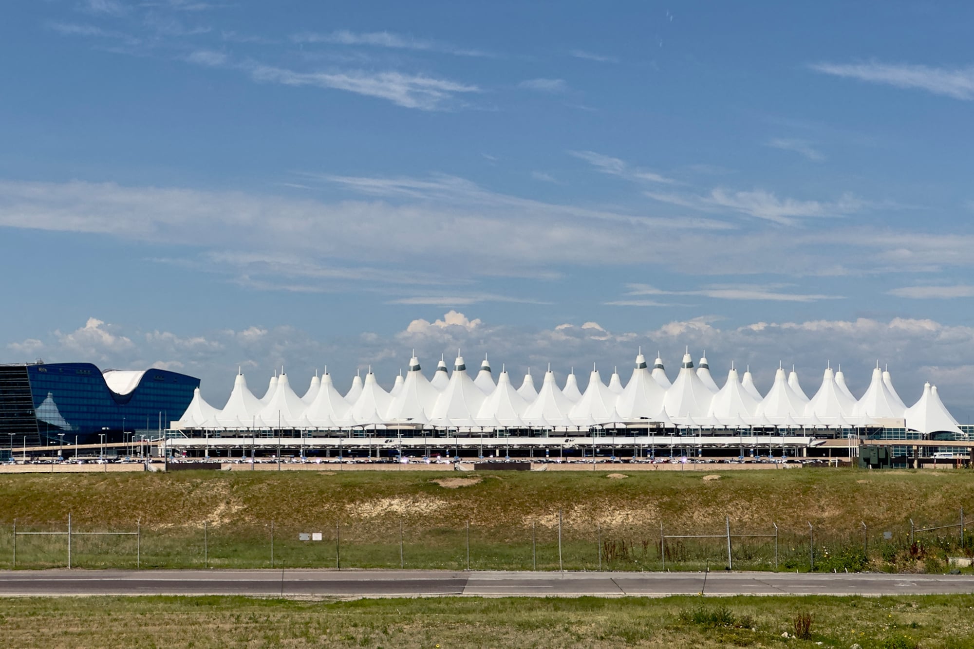 A photograph of Denver International Airport, which is a large series of white coned roof with a big blue sky with some clouds and a grassy field in the foreground.
