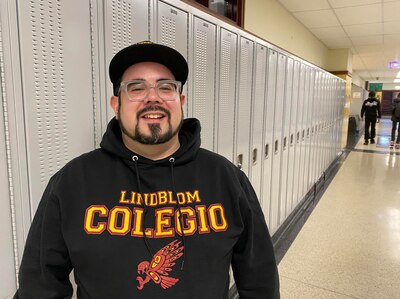 A man wearing a dark sweater and a black hat poses for a portrait in front of a row of lockers with students walking in the background.