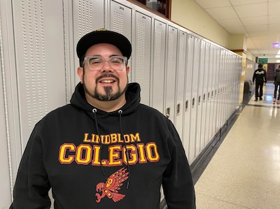 A man wearing a dark sweater and a black hat poses for a portrait in front of a row of lockers with students walking in the background.