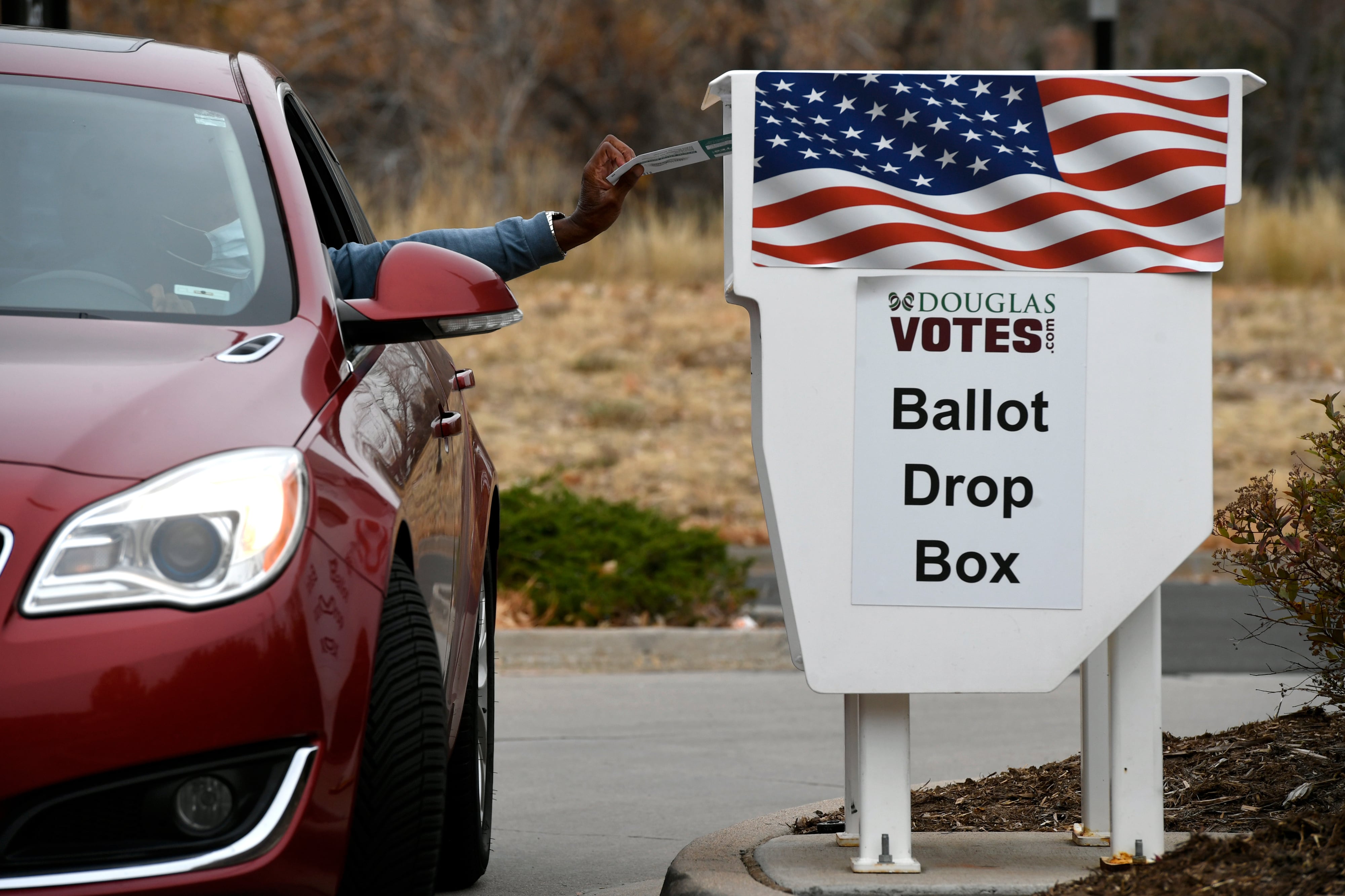A driver in a red sedan drops a ballot into a box that reads “McDouglas VOTES. Ballot Drop Box.”