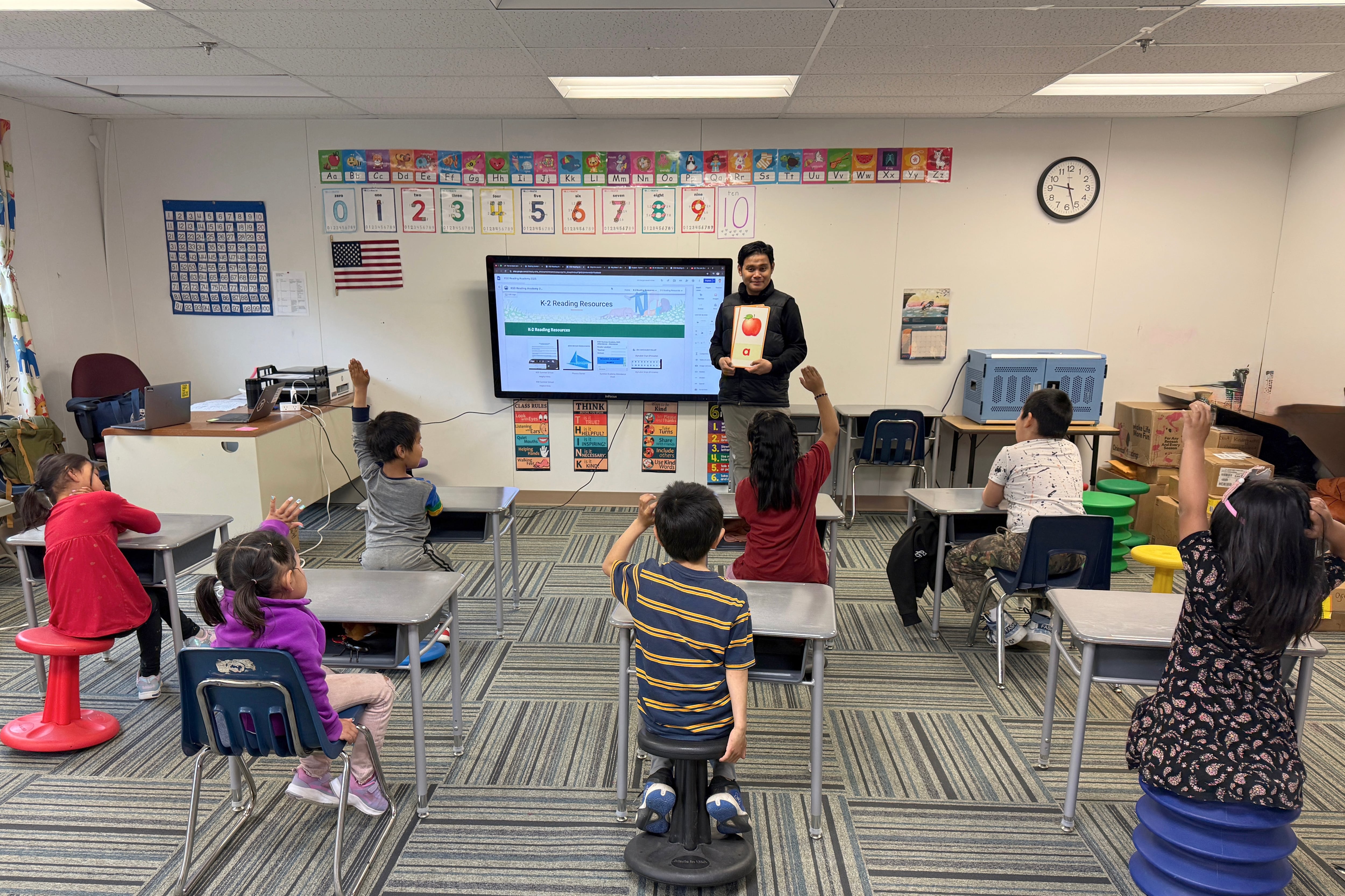A photograph of a man holding a giant letter card standing in front of a classroom with seven young children sitting in desks scattered across the room.