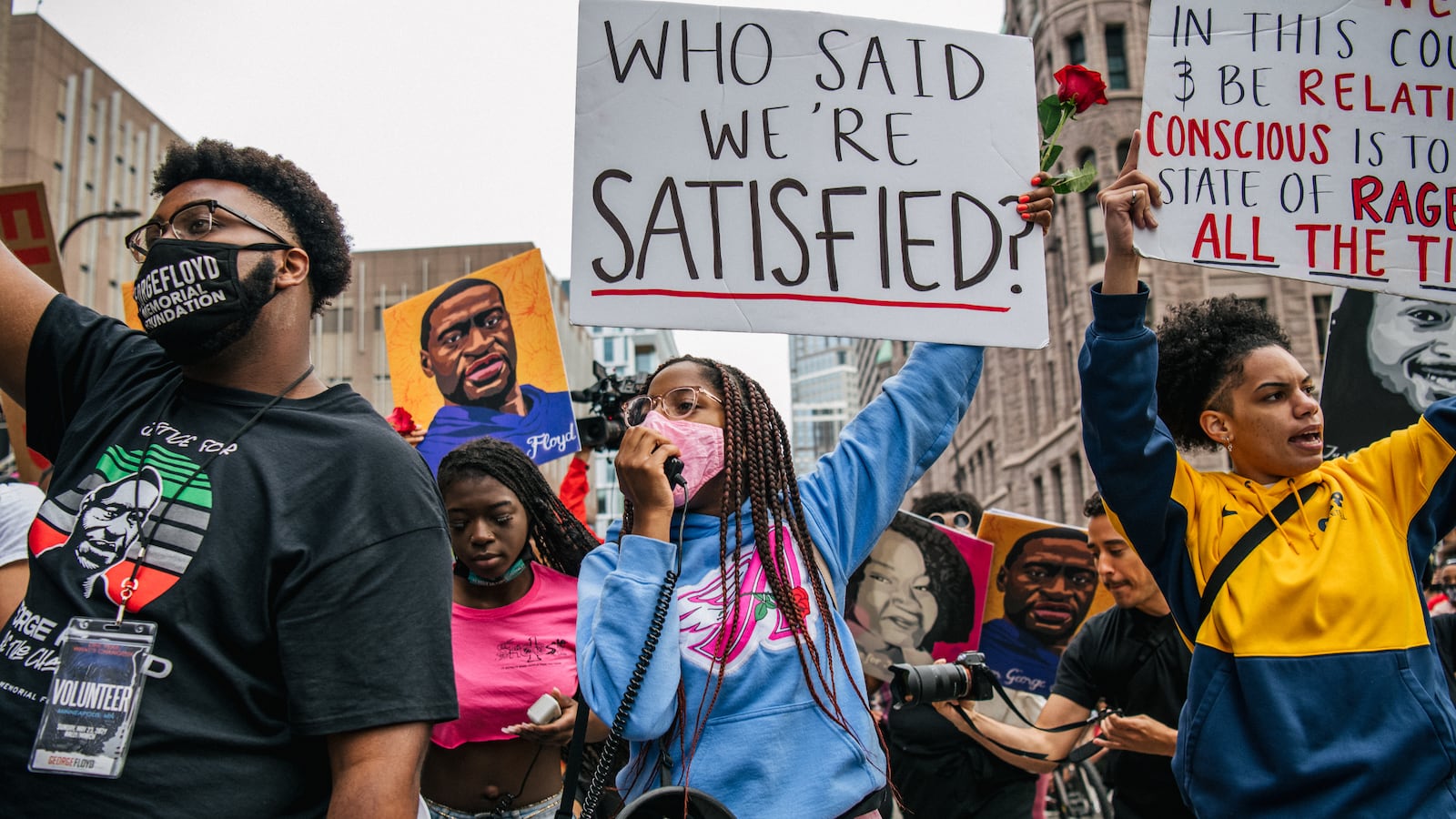 Demonstrators march in memory of George Floyd. In the center, a woman wearing a light blue hoodie and pink pants holds a sign that reads “Who said we’re satisfied?” and speaks through a bull-horn microphone.