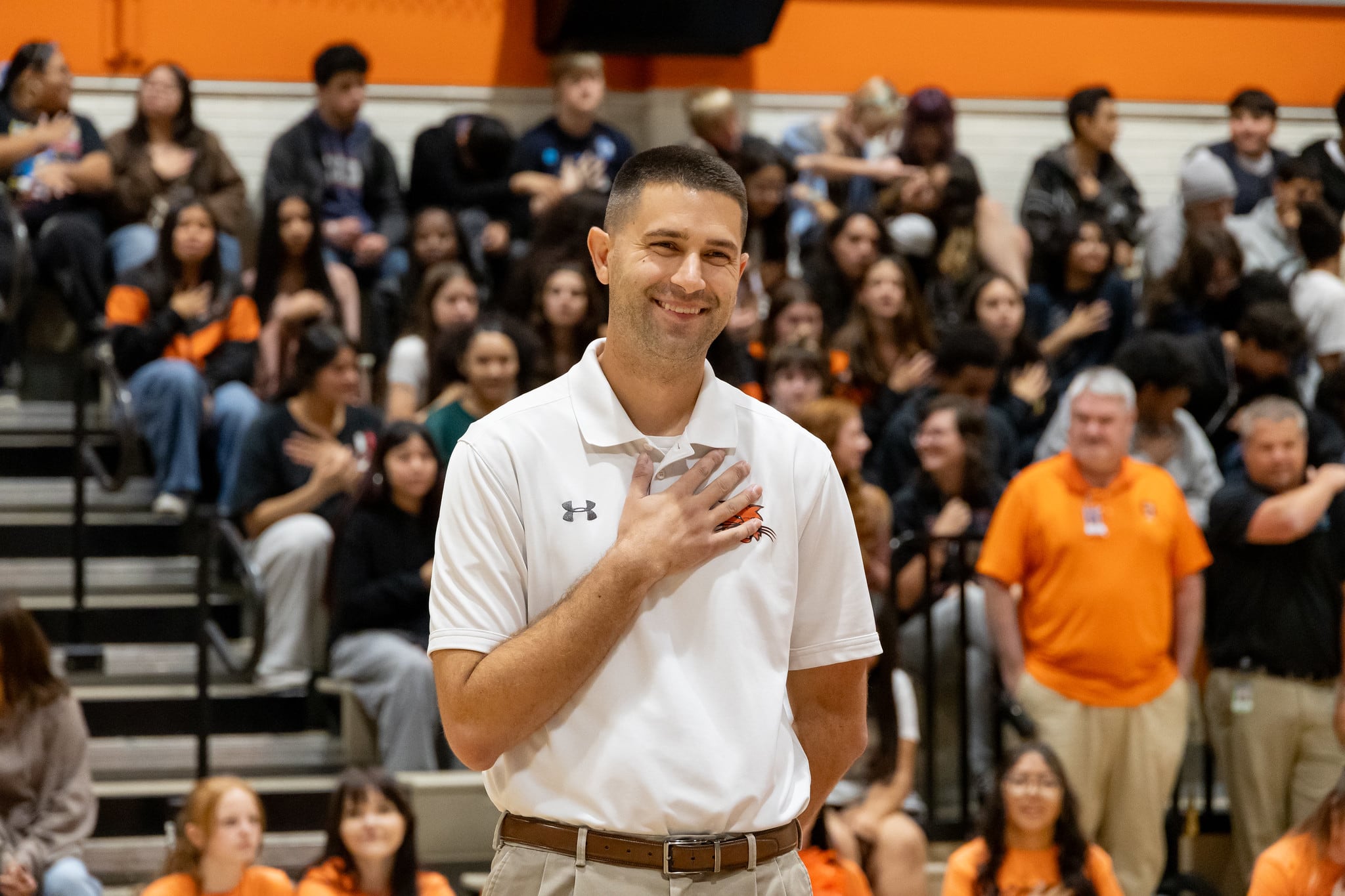 A photograph of a white man in a white shirt with his hand over his heart showing gratitude with a school gym full of students and adults.