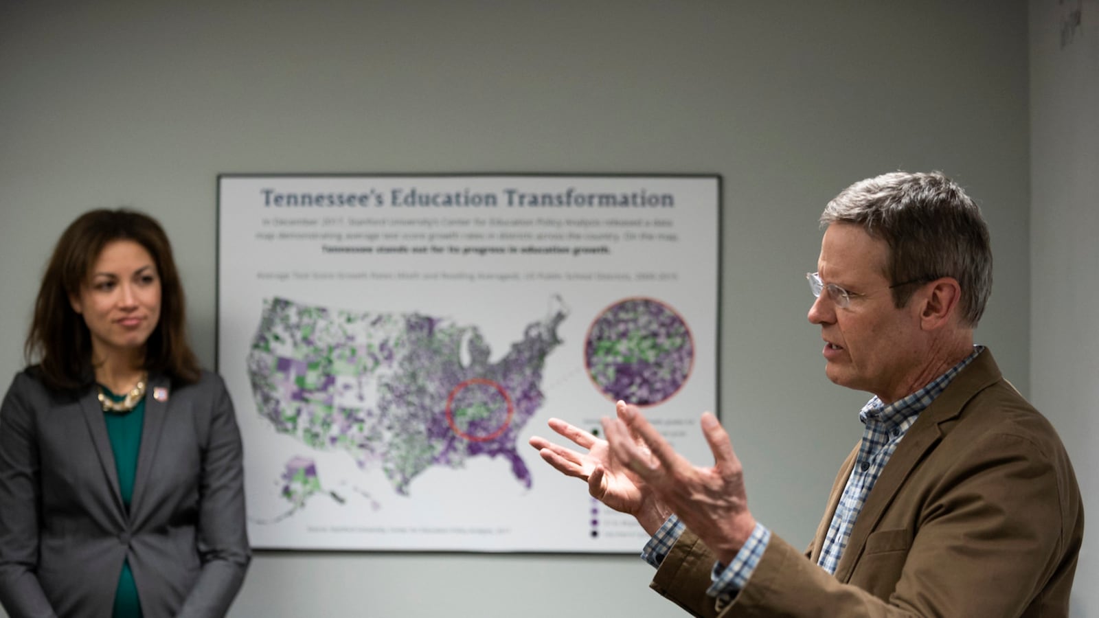 A man gestures while speaking, as a woman looks on in an office setting.