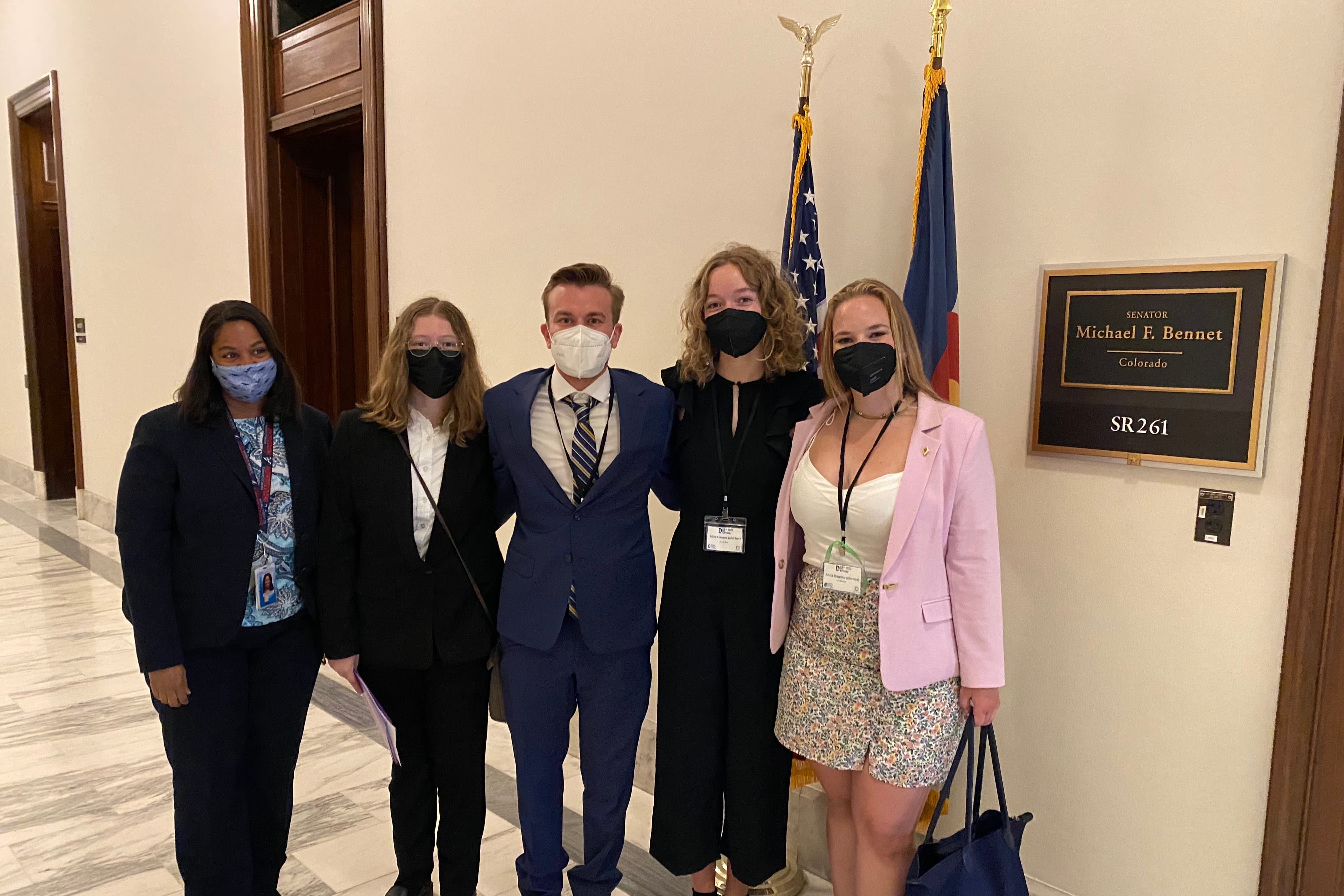 A group of young people in suit jackets and dress pants or skirts pose for a photo in the hallway of a Congressional office building. They’re wearing masks and standing shoulder to shoulder. A sign indicates they are in front of the office of U.S. Senator Michael Bennet.