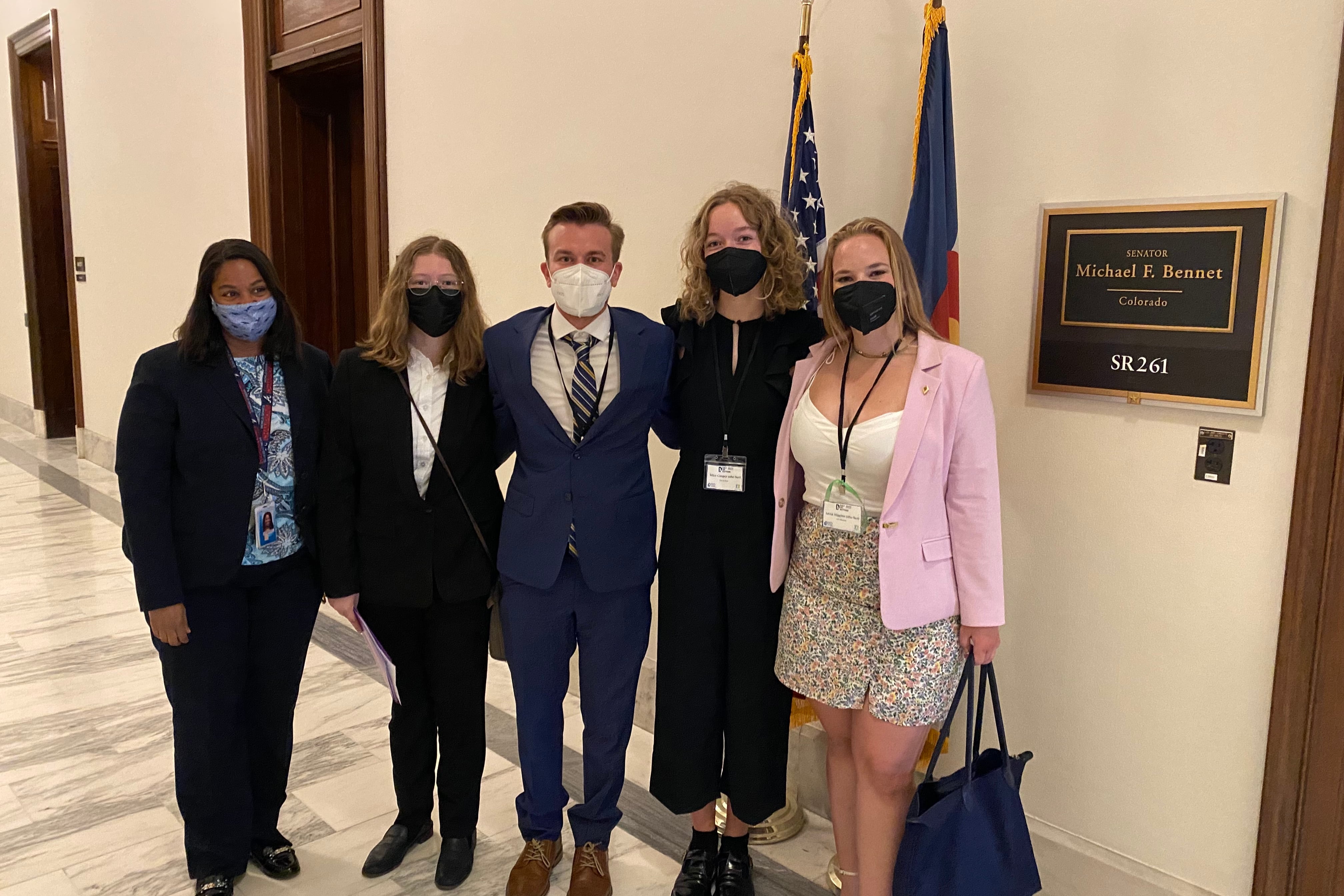 A group of young people in suit jackets and dress pants or skirts pose for a photo in the hallway of a Congressional office building. They’re wearing masks and standing shoulder to shoulder. A sign indicates they are in front of the office of U.S. Senator Michael Bennet. 