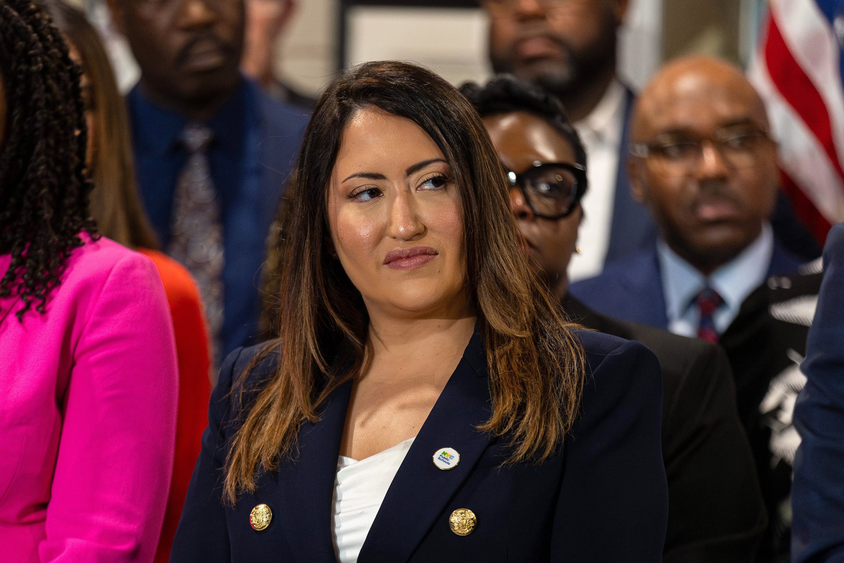 A photograph of a woman with long dark hair in a suit looking to the side while she stands in a crowd of people.