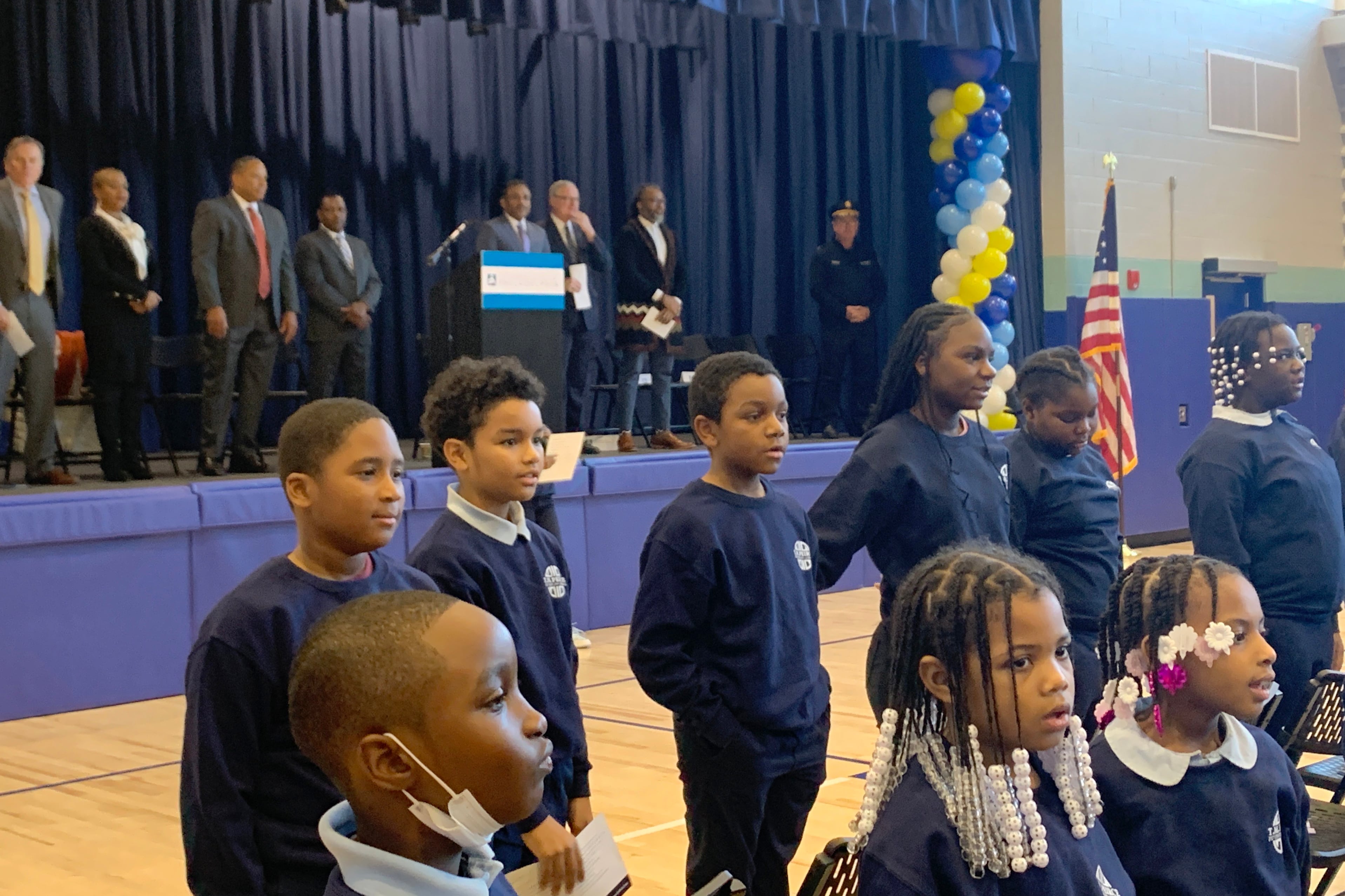 Students and adults stand on a stage in a school gym and on the gym floor.