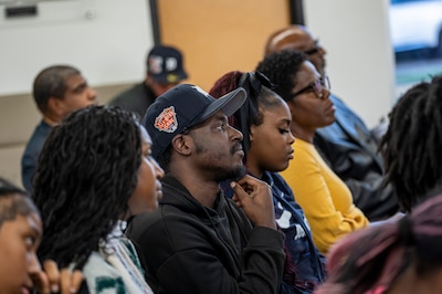 Several people sit in an audience and appear to be listening intently as candidates for the Detroit school board speak.