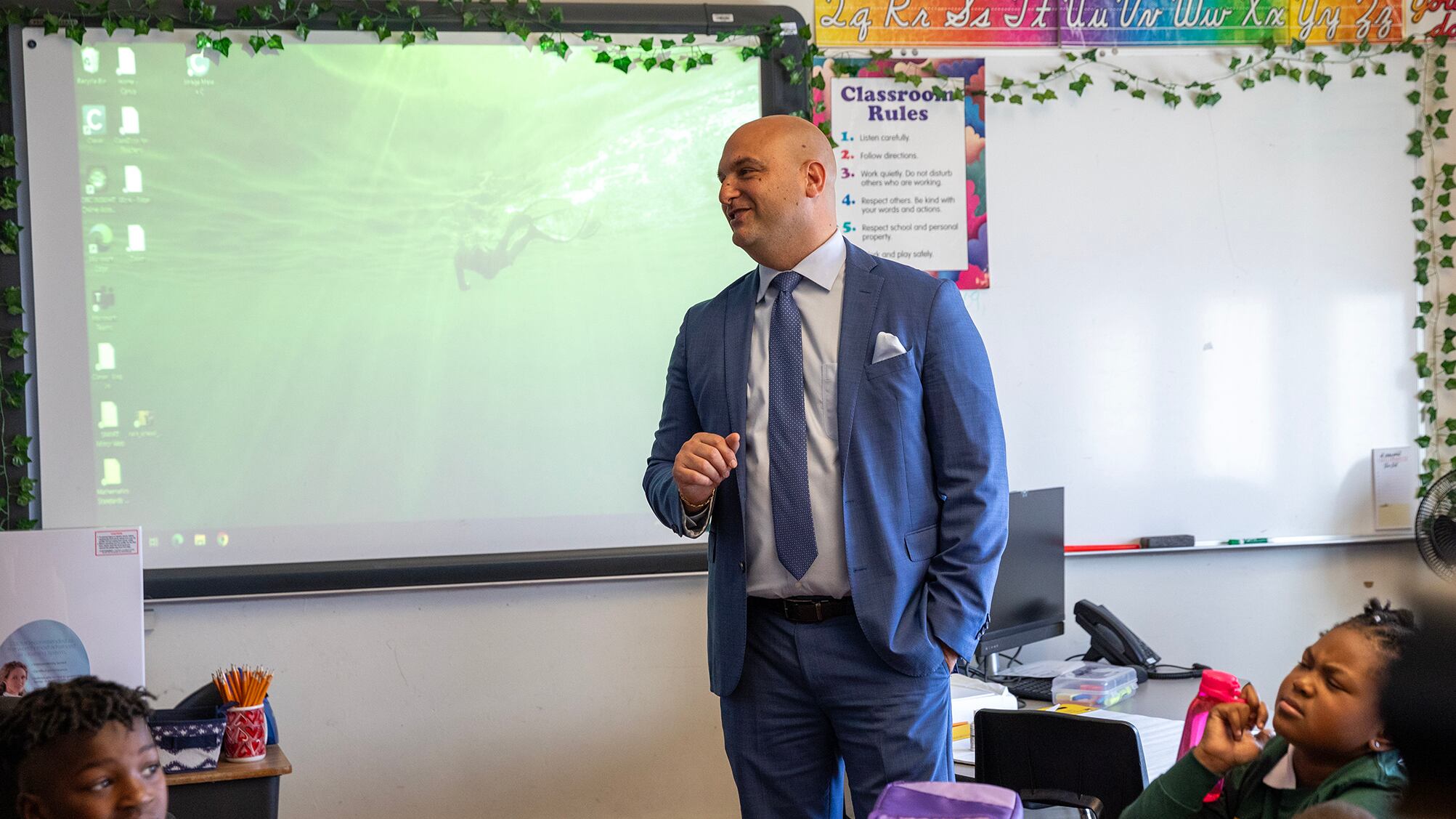 Man wearing a blue suit and tie at the front of a school classroom.