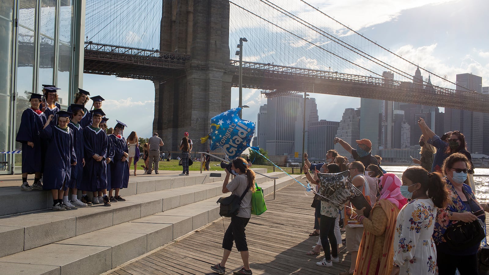 Students stand in graduation robes and caps next to the river and Brooklyn Bridge as a photographer takes photos