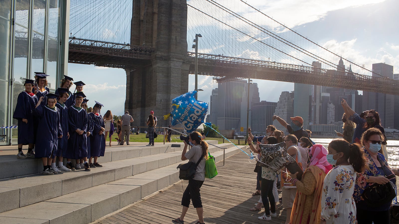 Students stand in graduation robes and caps next to the river and Brooklyn Bridge as a photographer takes photos