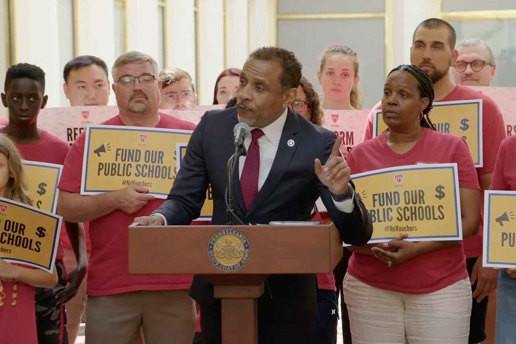 A man in a dark suit stands in front of people holding signs.