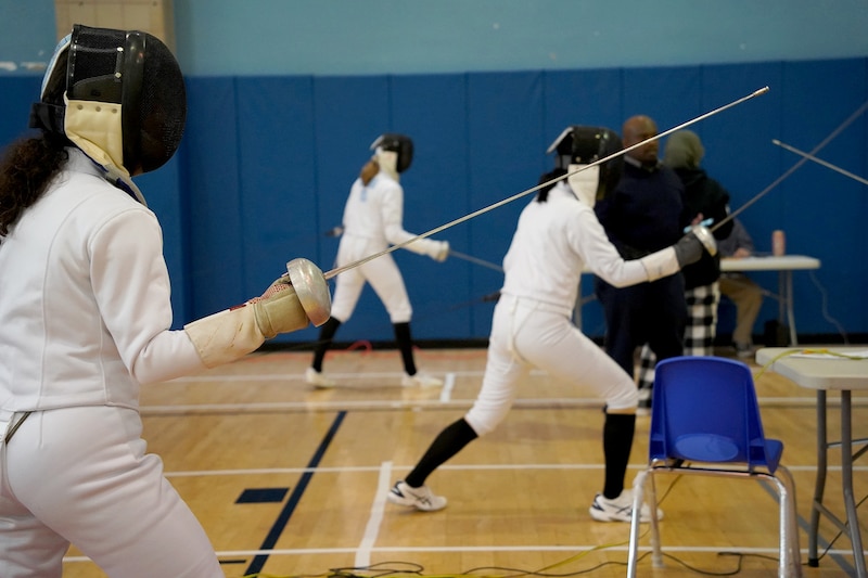 A student with long hair and dressed in an all white fencing uniform holds a fencing sword while two other students are in the background.