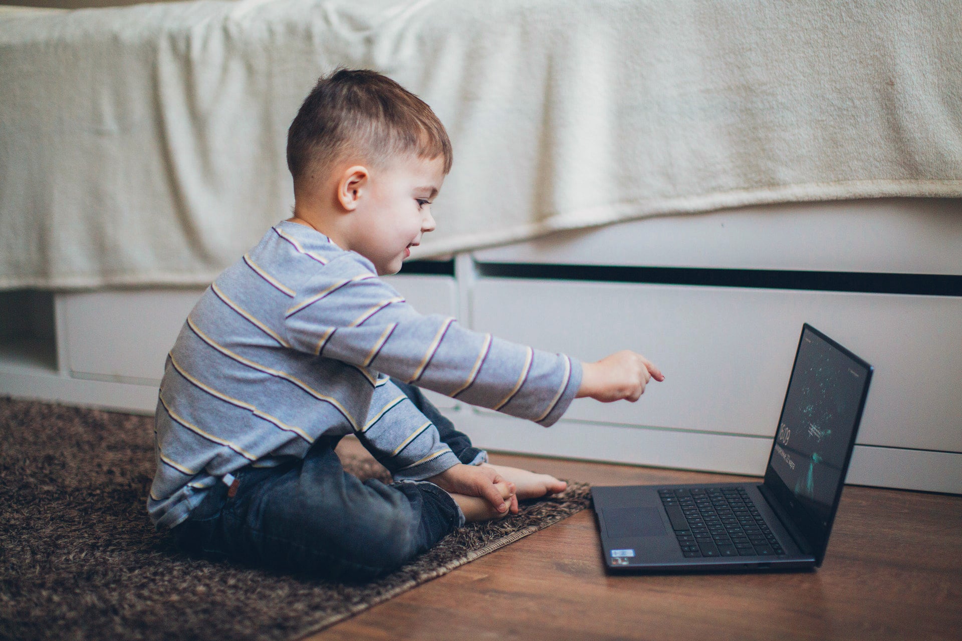 Young boy sitting on a rug pointing at a laptop screen.
