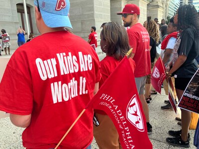A photograph of a group of people standing outside listening to a press conference while wearing red t-shirts that read "Our Kids Are Worth It."
