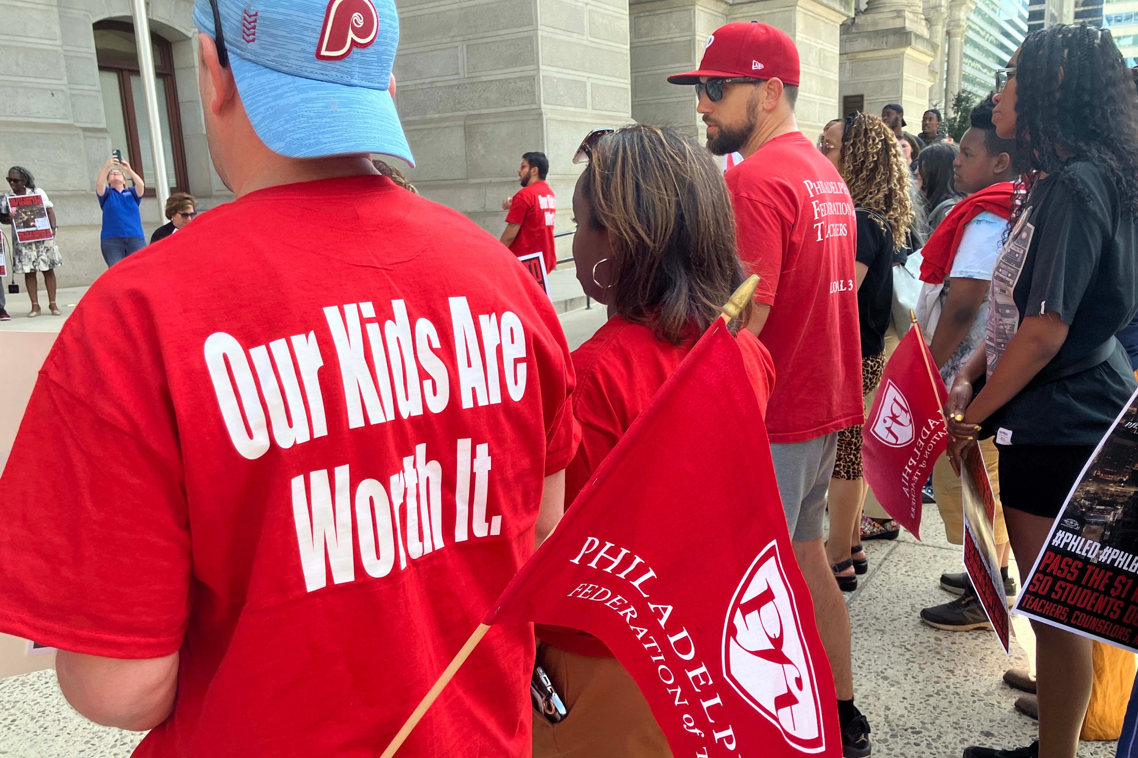 A photograph of a group of people standing outside listening to a press conference while wearing red t-shirts that read "Our Kids Are Worth It."