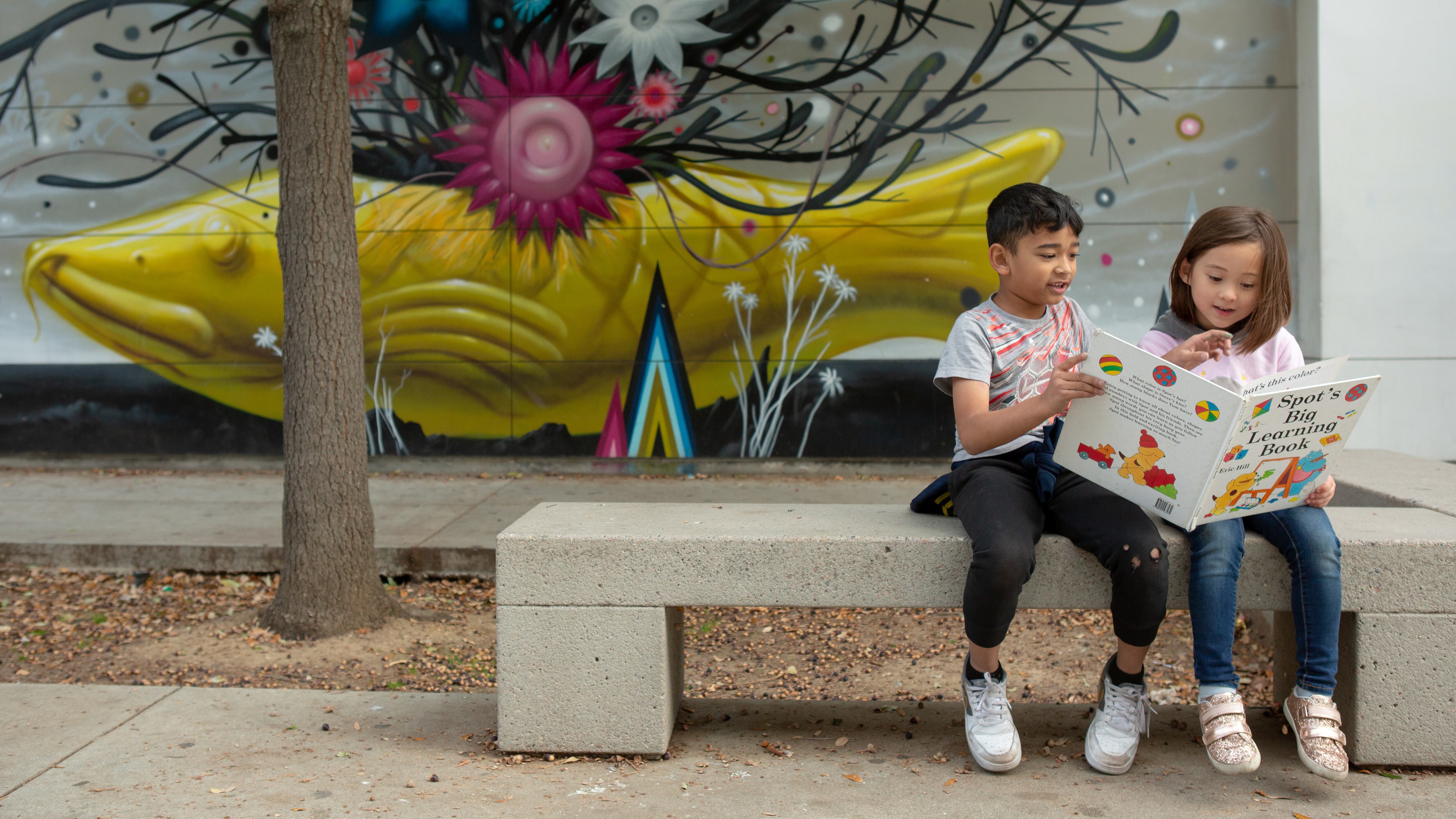 A boy and a girl share a picture book while sitting on a concrete bench in front of a concrete wall and a sculpture of a large yellow fish with a tangle of dark seaweed growing up from its back with blue, magenta and white flowers interspersed.