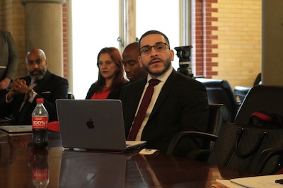 A man wearing a dark suit and wearing glasses sits at a wooden table with a laptop in front of him while he speaks. There are other people sitting behind him.