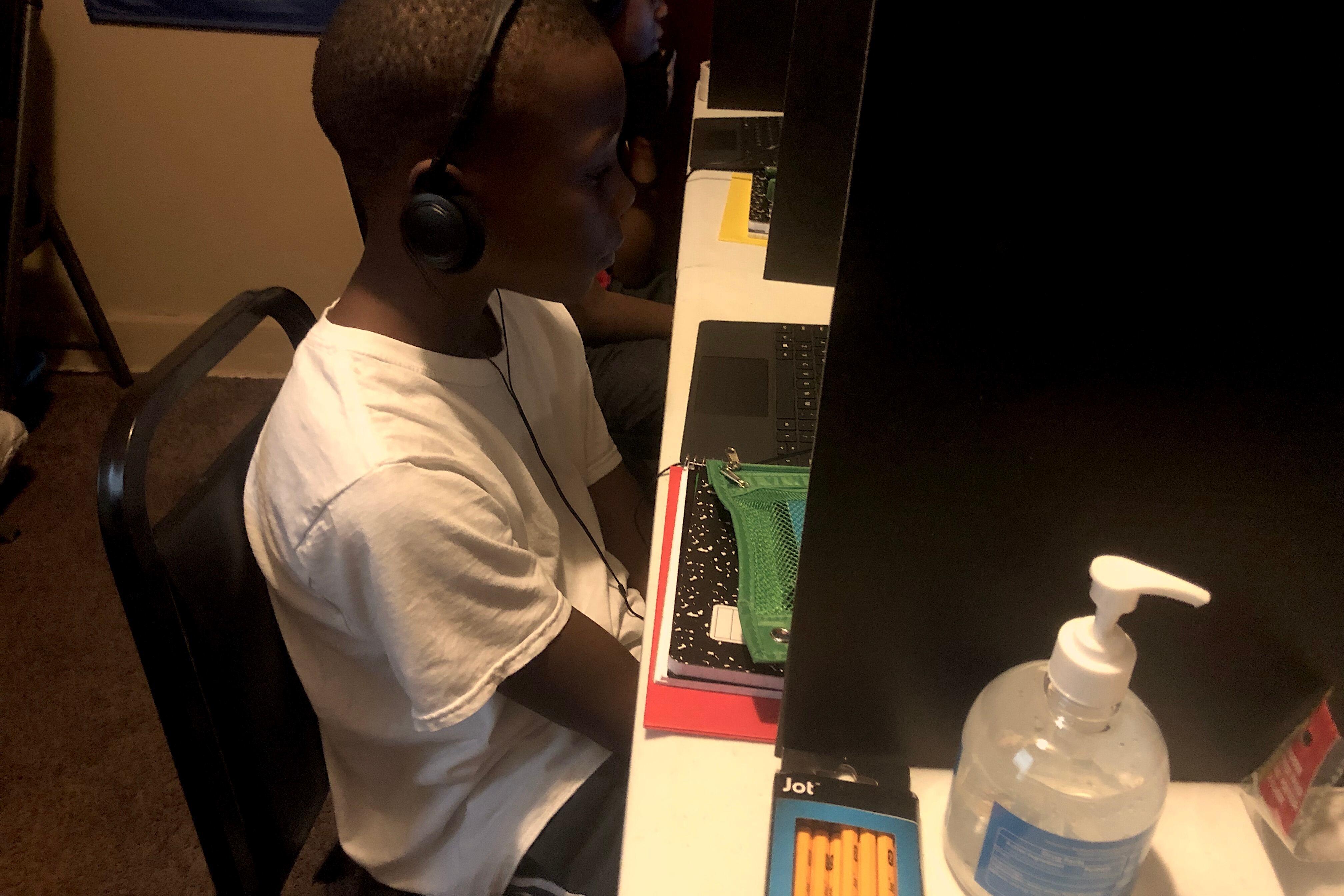 A third-grade student sits at a desk in front of a laptop with his hands in his lap as he listens to an online class.