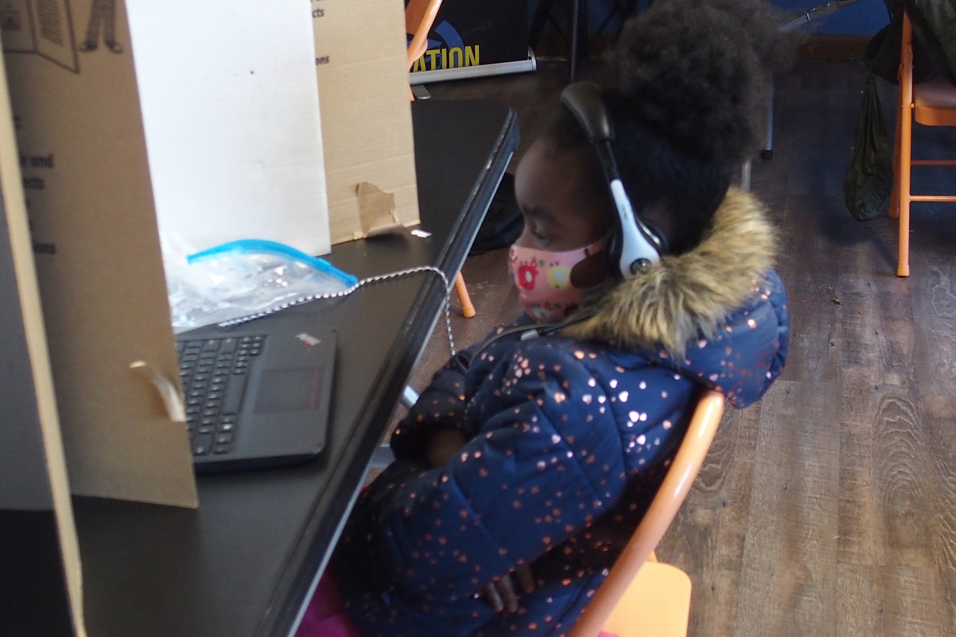A student in a coat and mask sits with her arms crossed in a metal folding chair at a table with a cardboard divider as she looks at her laptop for her online class.