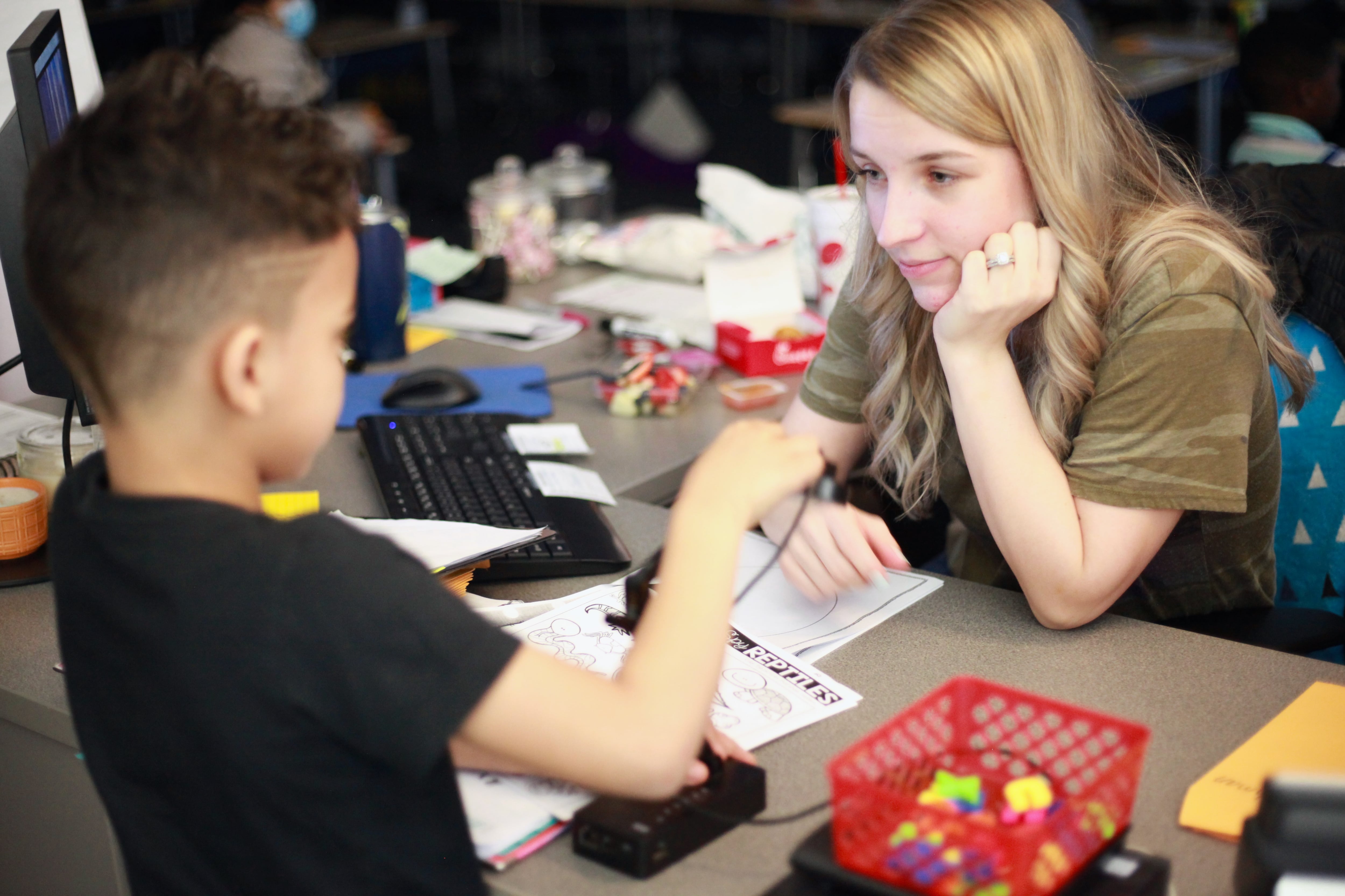 A young teacher works with a boy at her desk, who is motioning to her as she rests her hand on her chin.