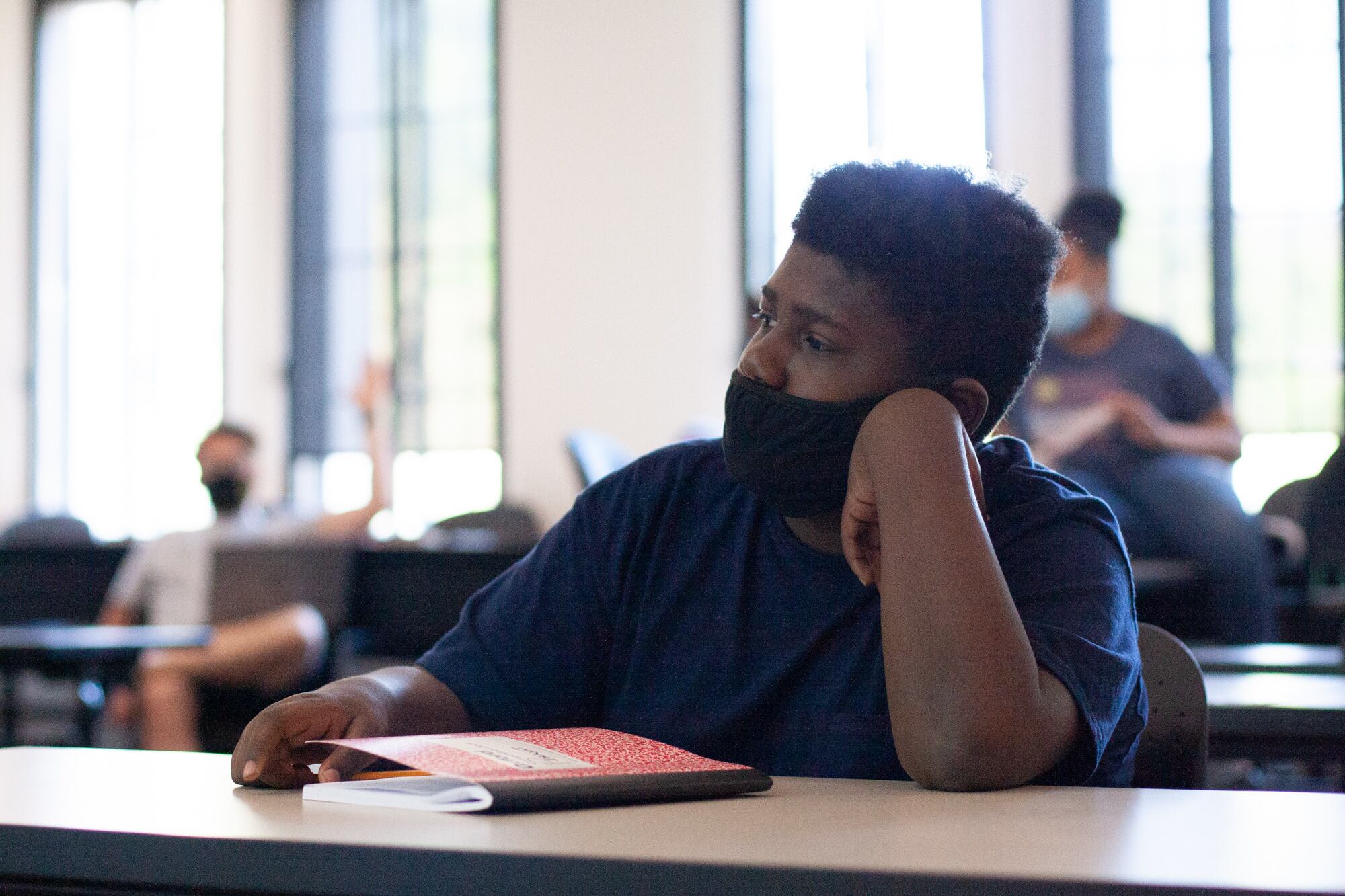 A sixth grade boy listens to a lecture, wearing a black mask and dark-colored shirt. He has a red composition notebook in front of him on the desk, and two other students sit behind him.
