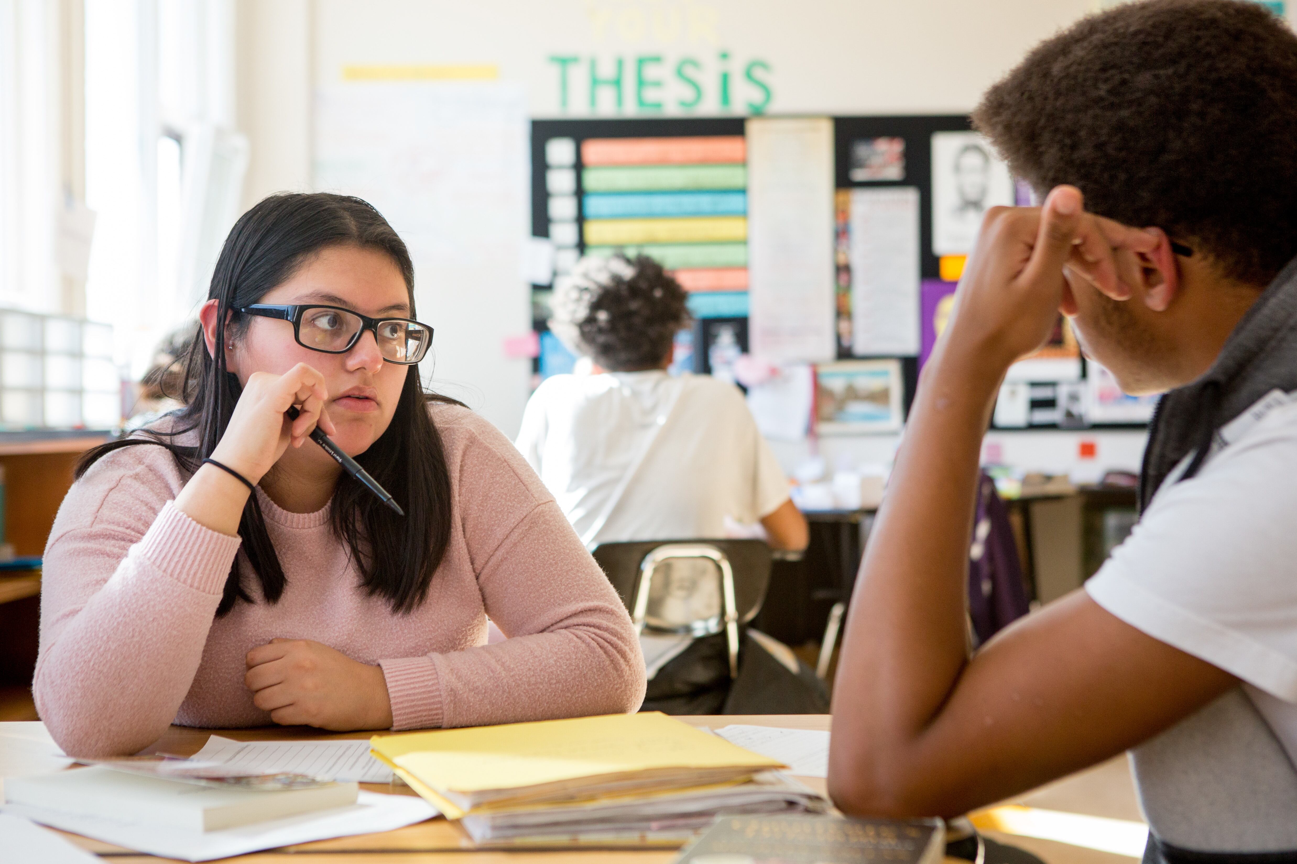 High school students look at each other mid-conversation as they sit at a table that has papers scattered all over.