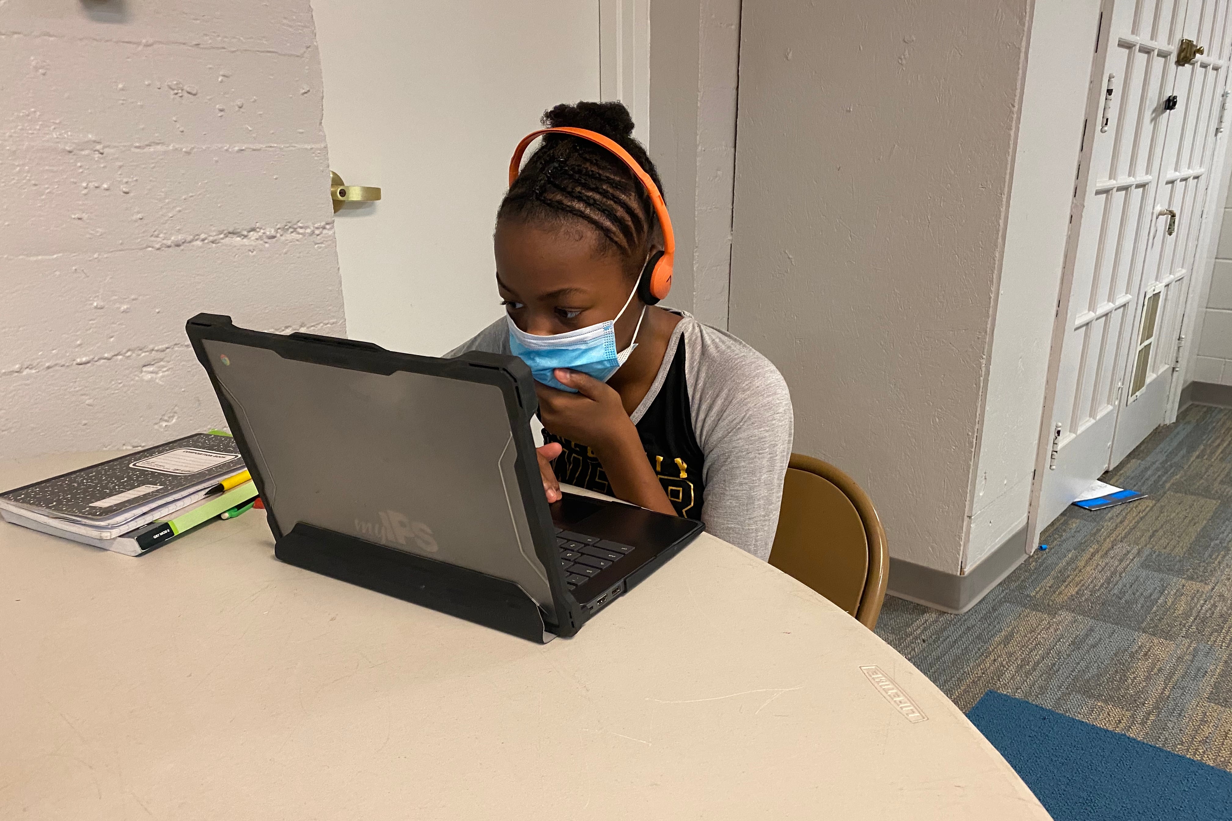 A student with a mask covering her face sits at a table looking at a laptop screen