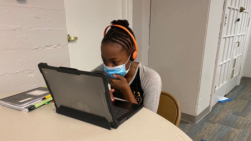 A student with a mask covering her face sits at a table looking at a laptop screen