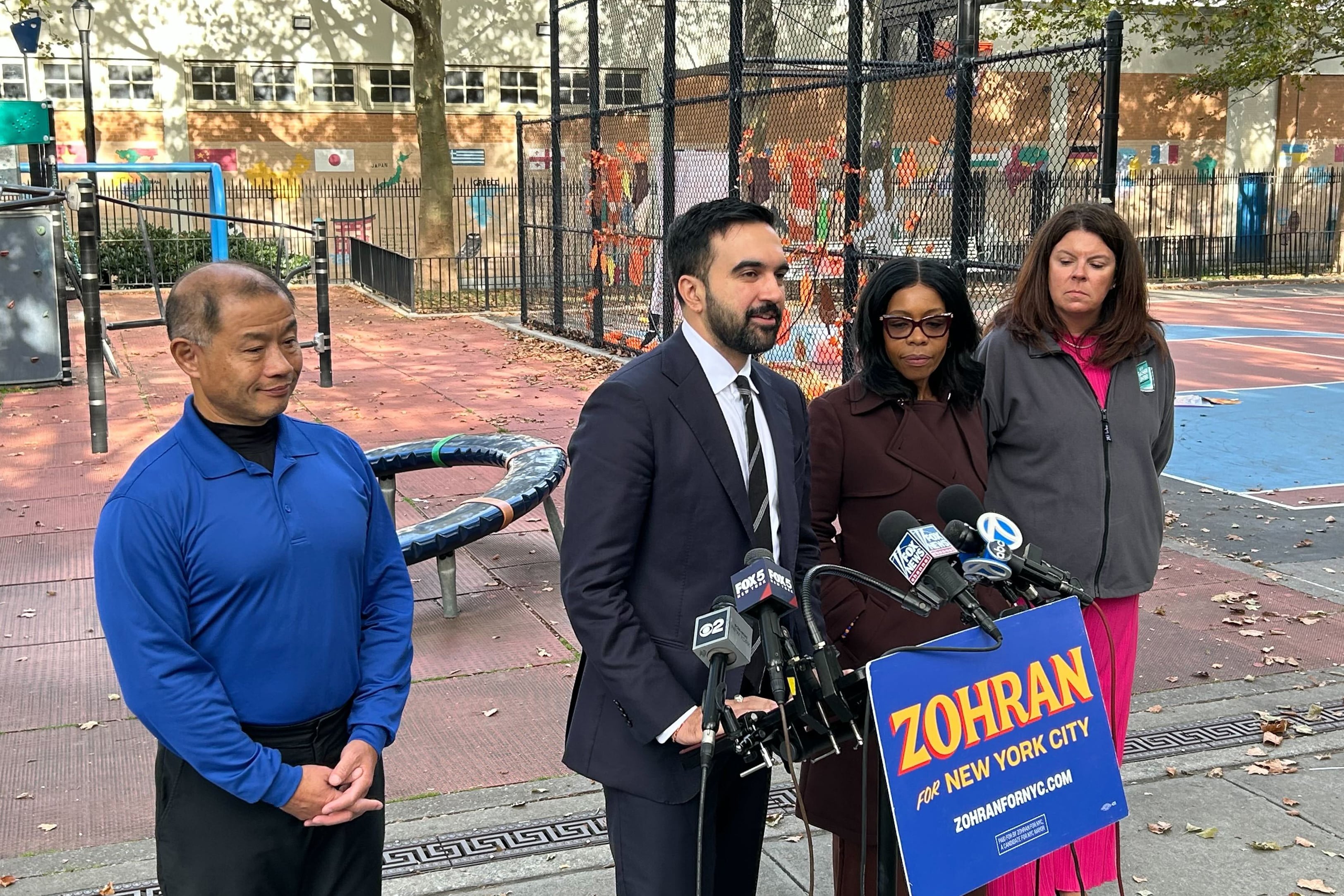 A photograph of four adults standing next to each other in front of a podium full of microphones and a sign that reads "Zohran for New York City" all in front of a school playground.