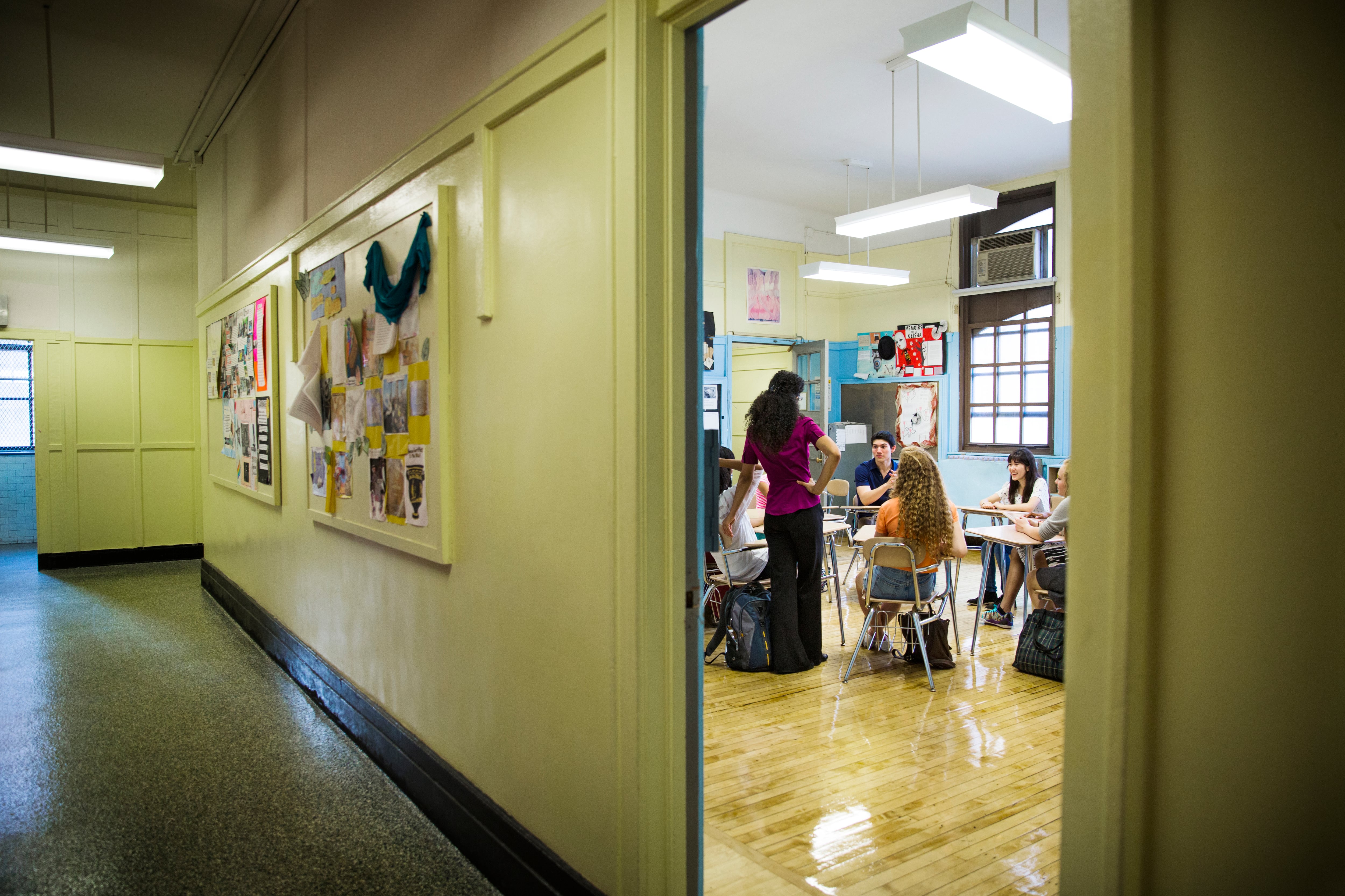 A view of students and a teacher in a classroom seen through a door and a hallway on the left.