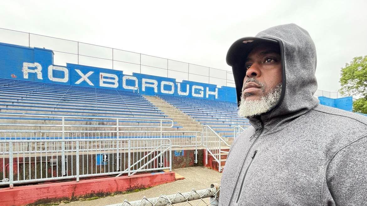 A man with a bear and a gray hooded sweatshirt stands in front of a chain link fence and a set of bleachers, with “Roxborough” on a blue wall behind the bleachers.