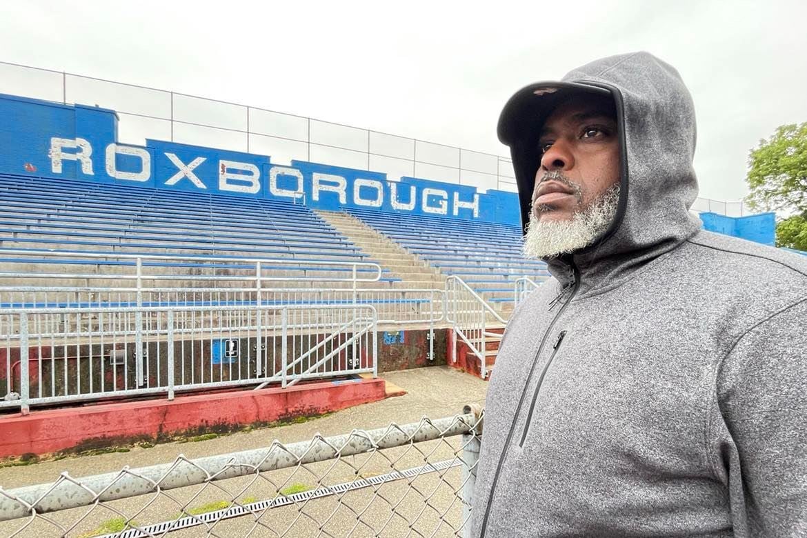 A man with a bear and a gray hooded sweatshirt stands in front of a chain link fence and a set of bleachers, with “Roxborough” on a blue wall behind the bleachers.