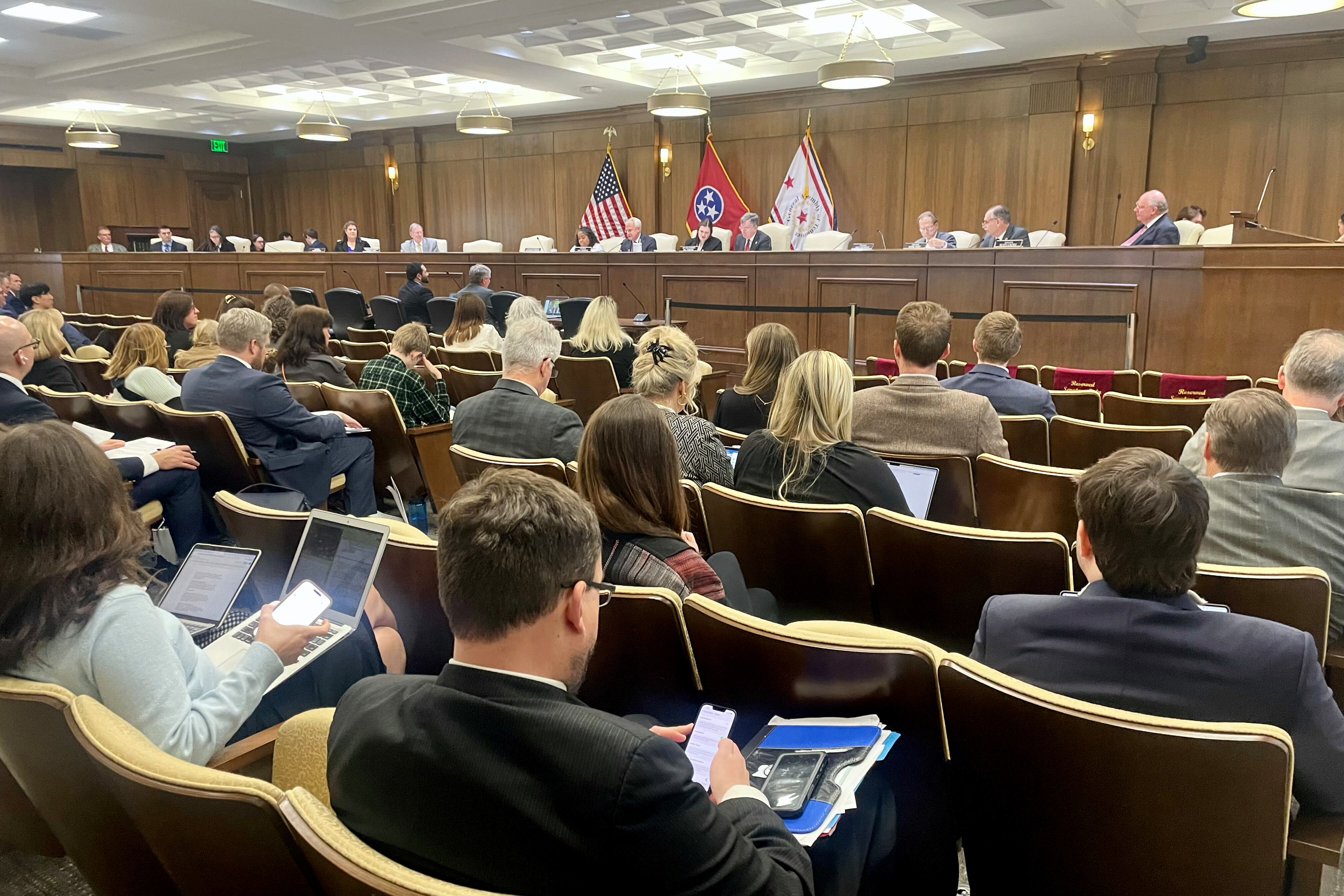 A room full of people sit in chairs in rows while a line of people sit at a wooden podium in the back.
