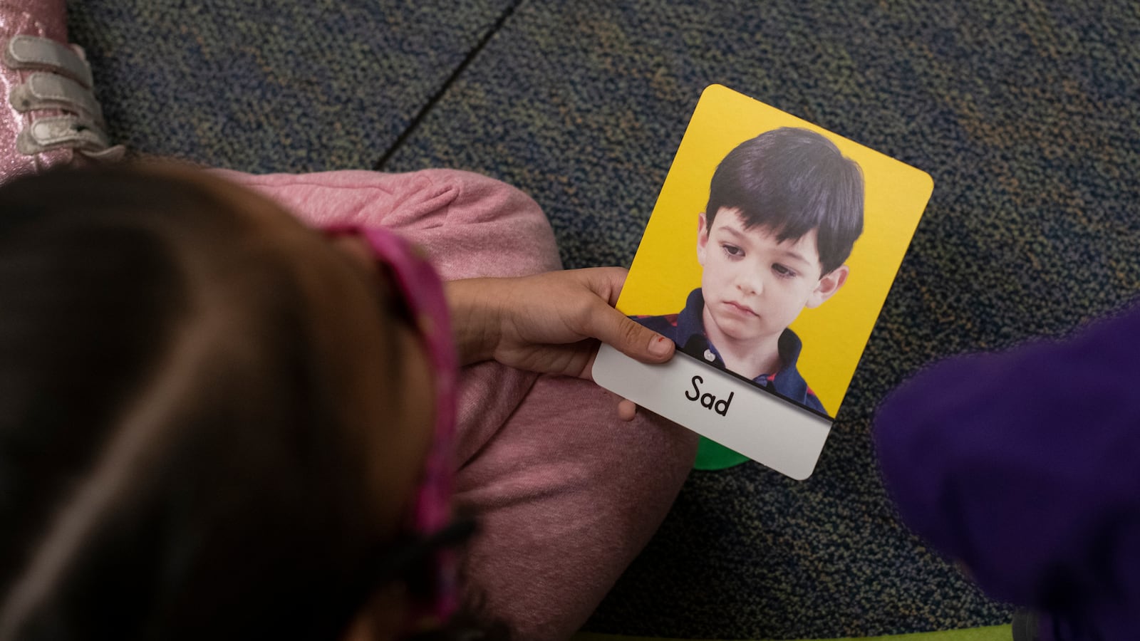 A girl in a pink dress holds a card with a boy’s sad face that says “sad.”