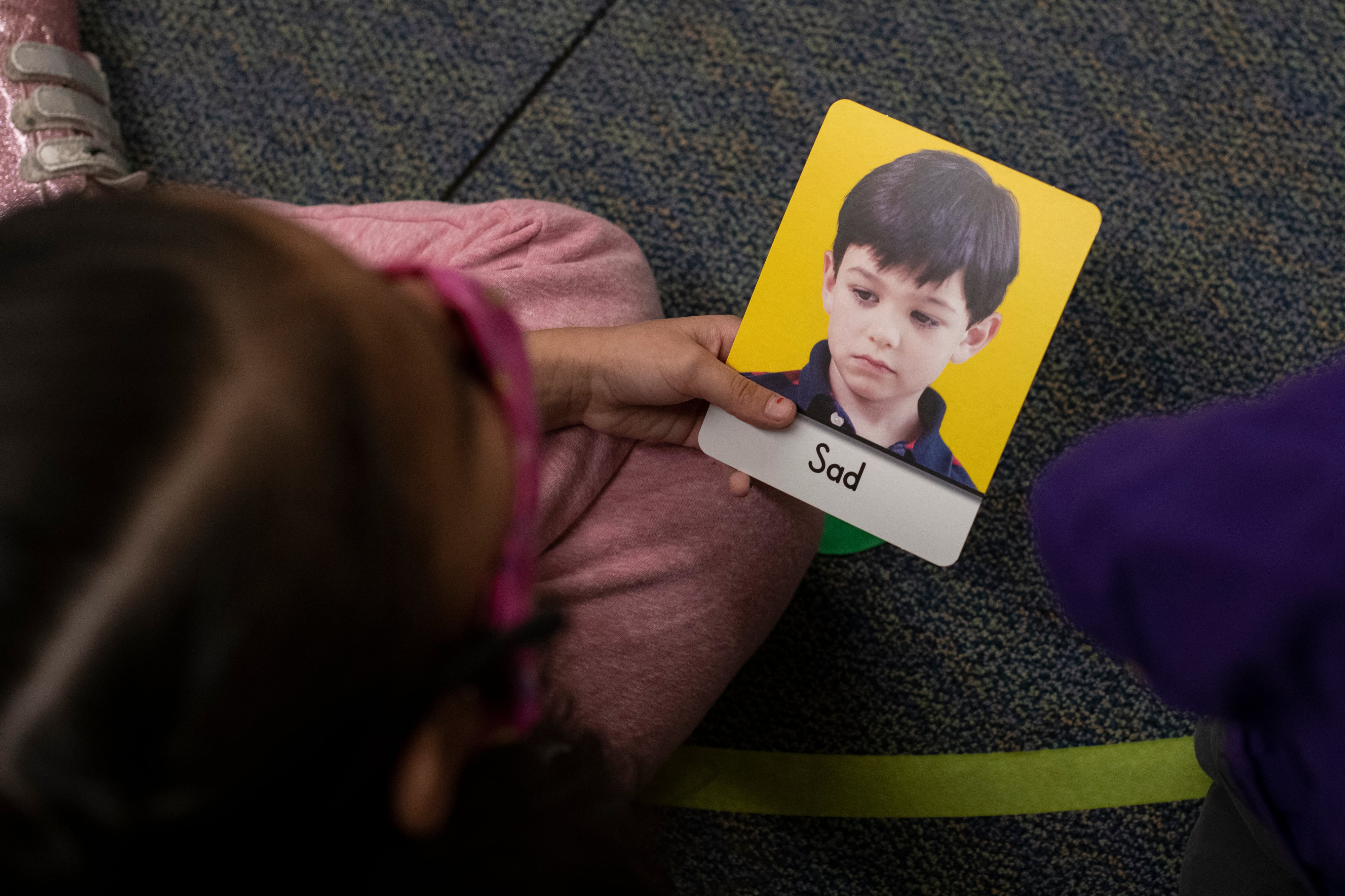 A girl in a pink dress holds a card with a boy’s sad face that says “sad.”