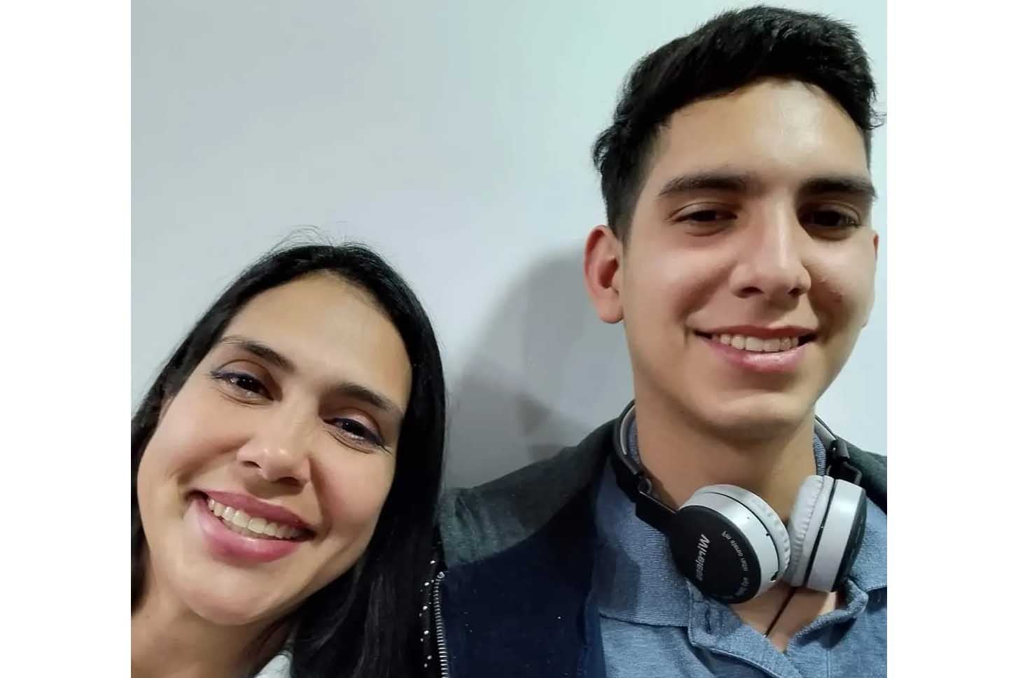 A photograph of two people with dark hair smile for a selfie against a wall.